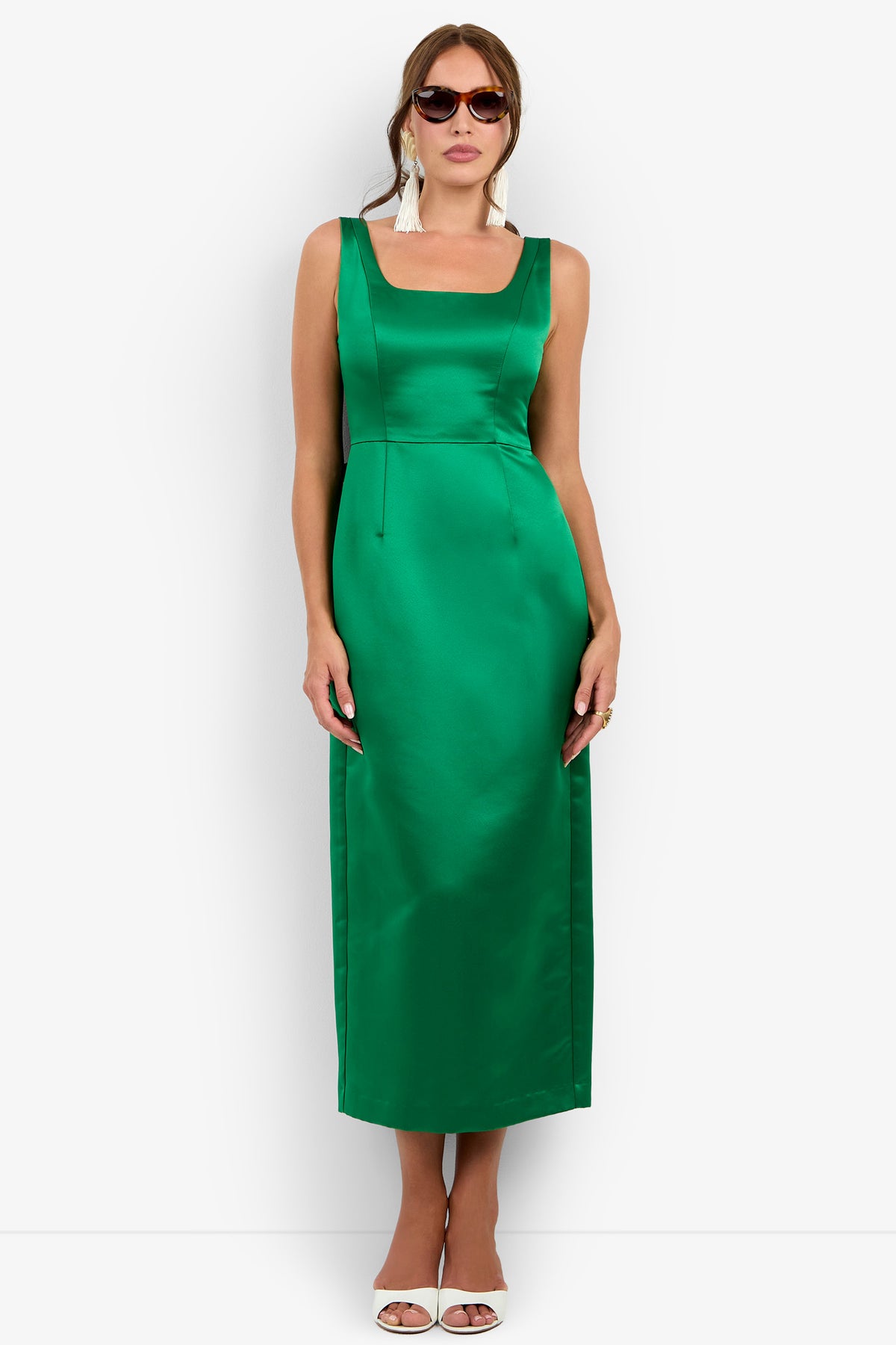 A woman wearing the Philia Dress—a sleeveless, emerald green satin dress with a square neckline—paired with white tassel earrings, sunglasses, and white heels, stands against a white background.