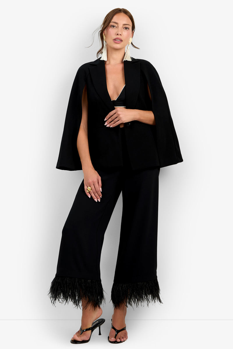 Wearing the Volterra Jacket styled as a black cape blazer, paired with wide-leg pants featuring feathered hems, black sandals, and gold jewelry, a woman stands against a plain white background.