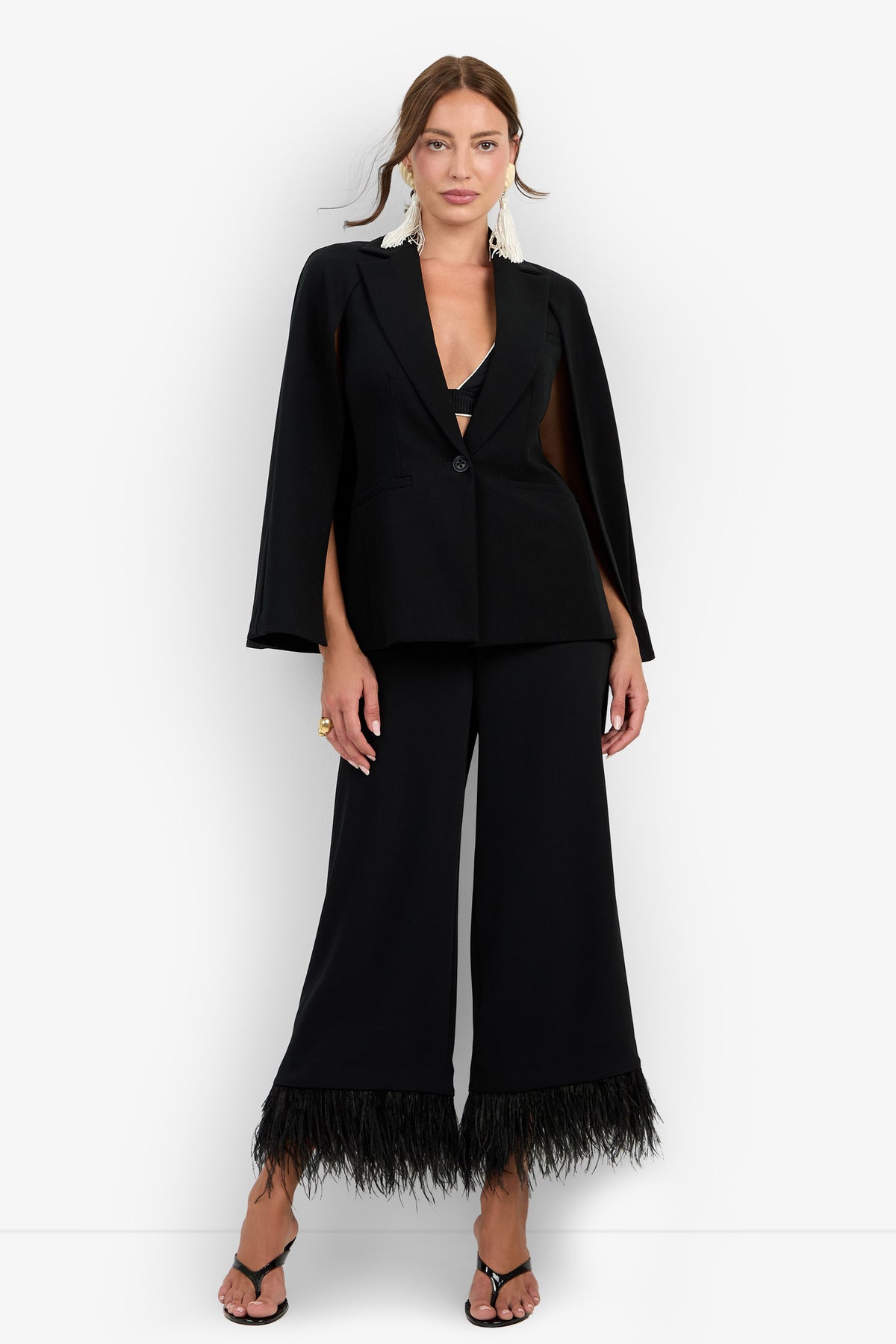 A woman stands against a plain white background wearing the Volterra Jacket in black—a cape-style blazer paired with wide-leg pants featuring feathered hems. She completes the look with black open-toe heels and large white statement earrings.