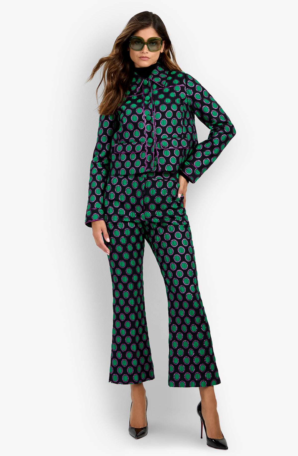 Woman wearing the Allex Jacket styled with matching flared pants featuring a geometric green and purple pattern, black high heels, and sunglasses, standing against a white background.