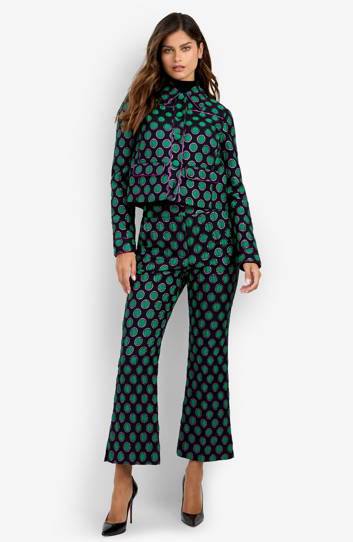 A woman standing against a white background wears the Allex Jacket—a cropped black jacket with green and purple circular patterns—paired with matching flared pants and black heels.