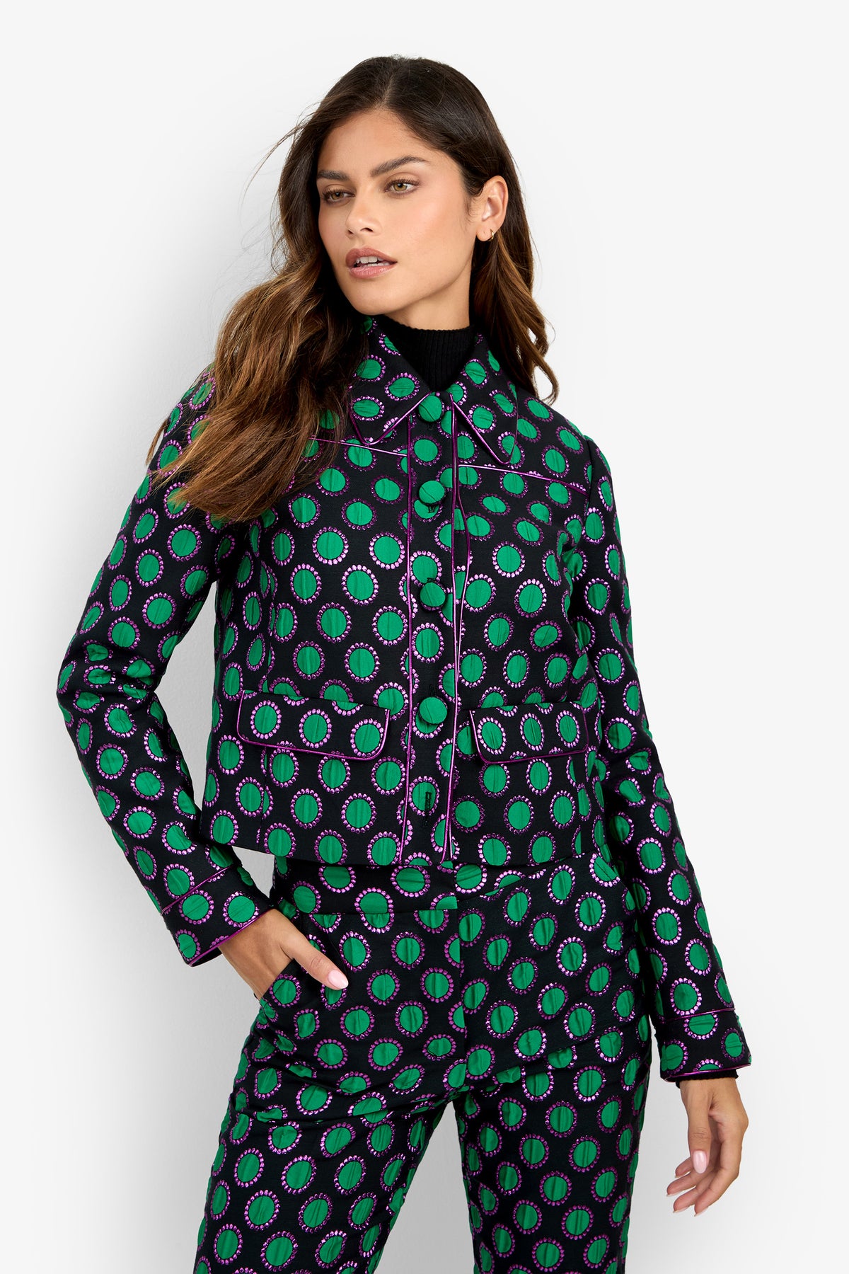 A woman poses against a plain white background wearing the Allex Jacket, featuring black, green, and purple circular patterns, paired with matching pants and a black turtleneck underneath.