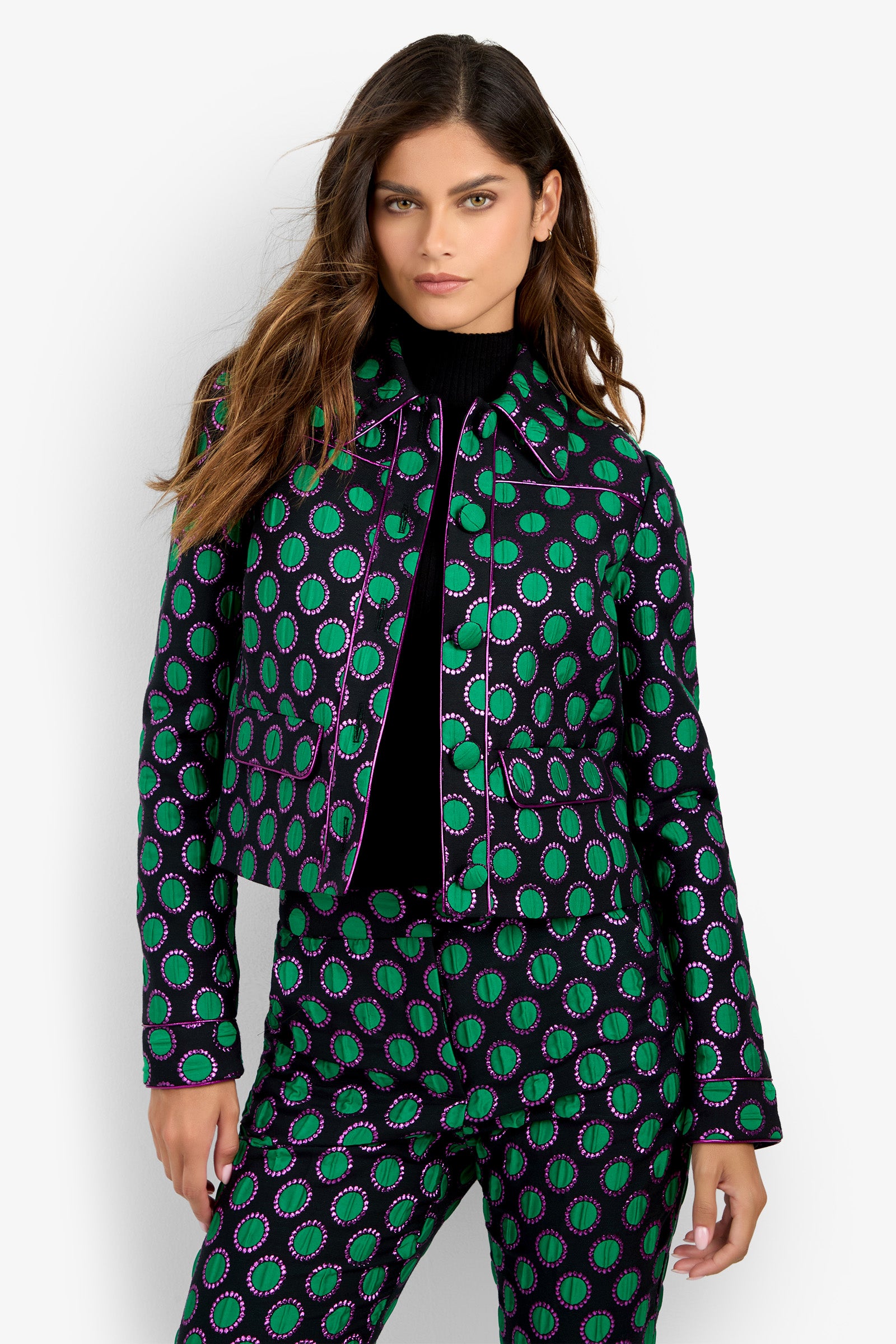 A woman with long brown hair wears the Allex Jacket—a black, green, and purple patterned design—paired with matching pants over a black turtleneck, standing against a plain white background.