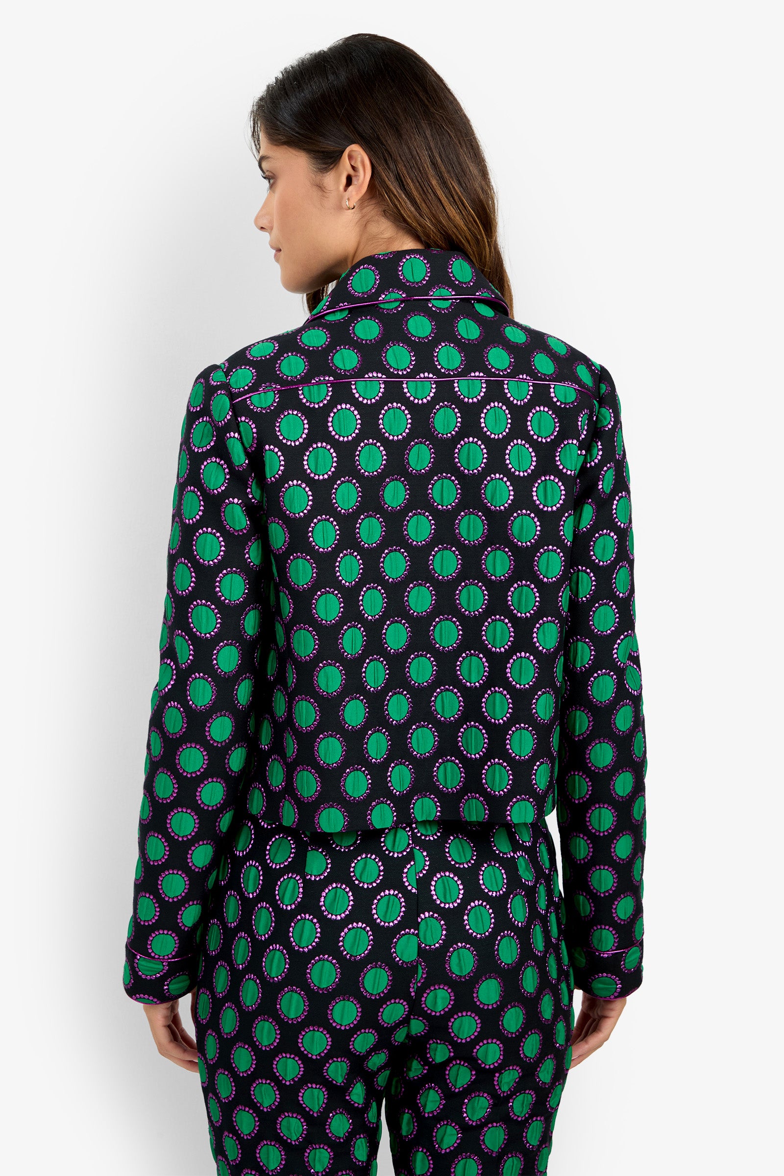 A woman faces away, wearing the Allex Jacket and matching pants featuring a vibrant green and purple circular pattern. Her brown hair is down, with a plain white background behind her.