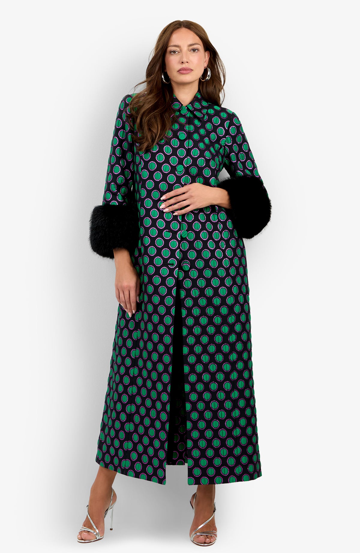 Dressed in the Dalaine Coat in MULTI with bold green and purple circles and black fur cuffs, the woman stands against a plain white background, one hand raised near her chest and paired with silver heels.