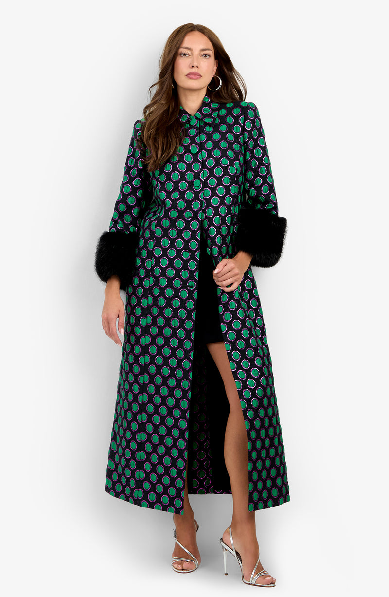 A woman wearing the Dalaine Coat, featuring green and blue circular patterns, black faux fur cuffs, and a high slit, paired with silver high-heeled sandals.