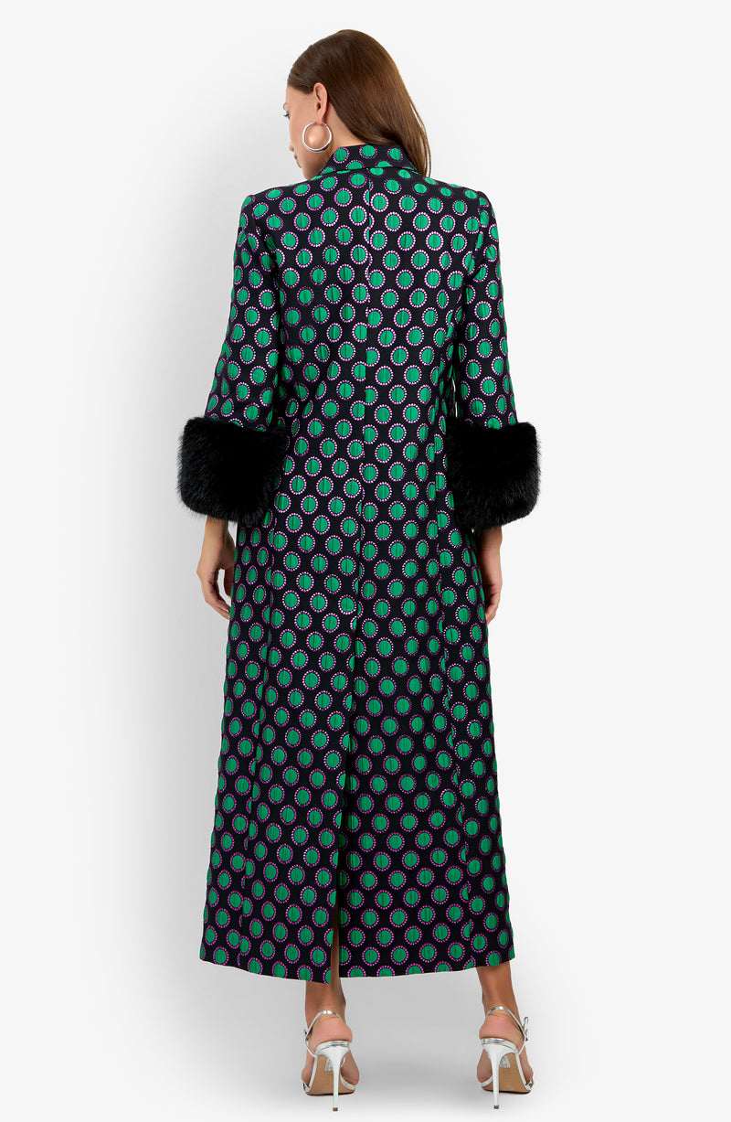 A woman, facing away, wears the Dalaine Coat—a long patterned piece featuring green and purple circles with black faux fur cuffs—paired with white heeled sandals and large hoop earrings against a white background.
