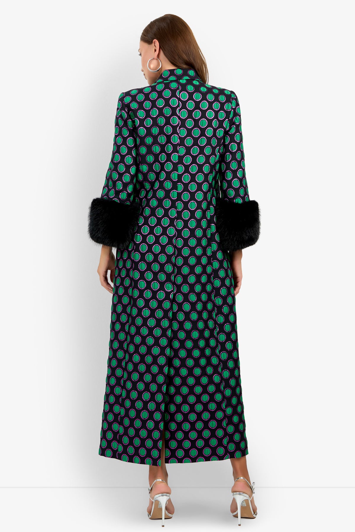 A woman stands with her back to the camera, wearing the Dalaine Coat—a long, patterned coat featuring green circles on a black background—styled with black faux fur cuffs, large hoop earrings, and white heeled sandals. The background is plain white.