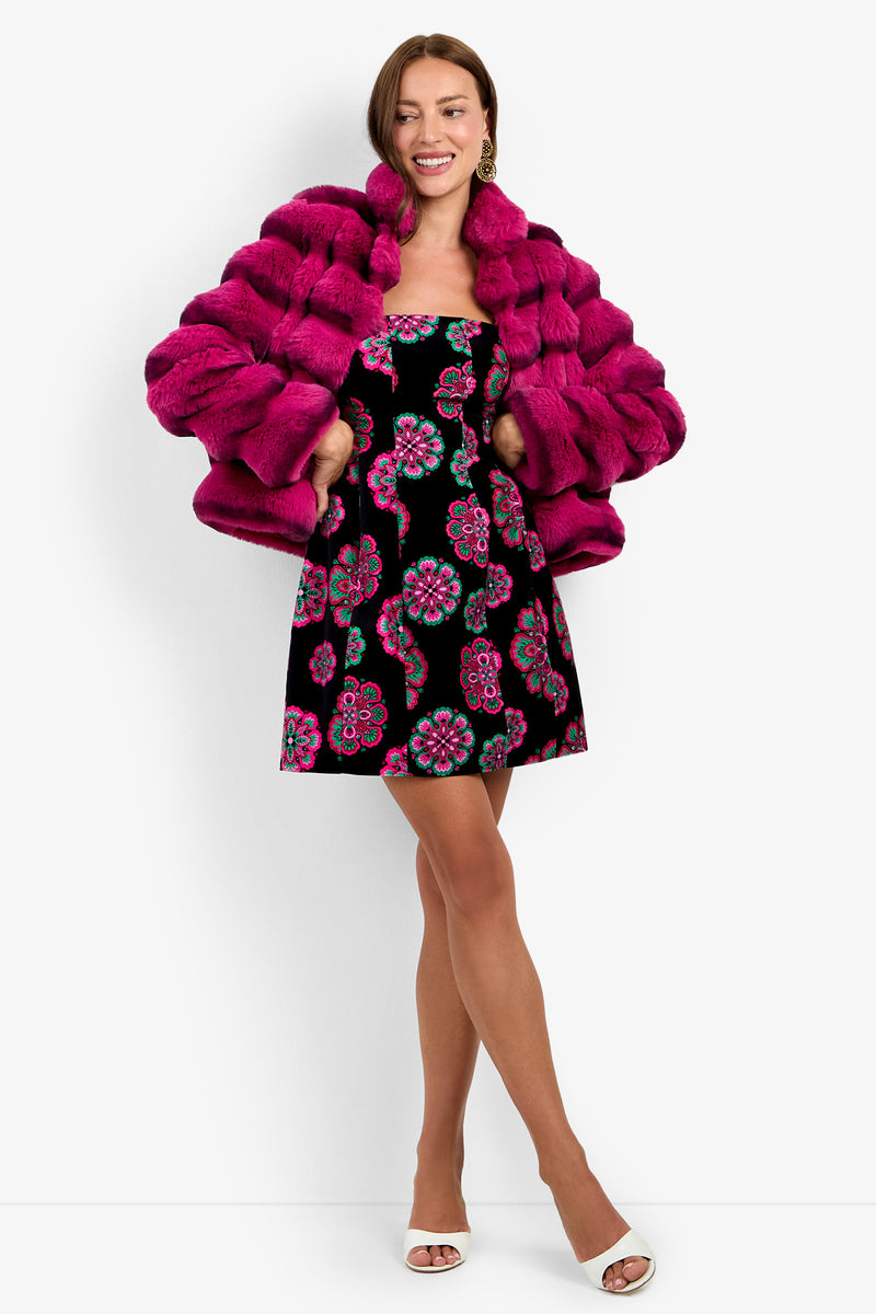 Smiling woman wearing the Mikel Coat in TORMALINA over a black dress with a colorful floral pattern, white open-toe heels, and hoop earrings, standing against a plain white background.