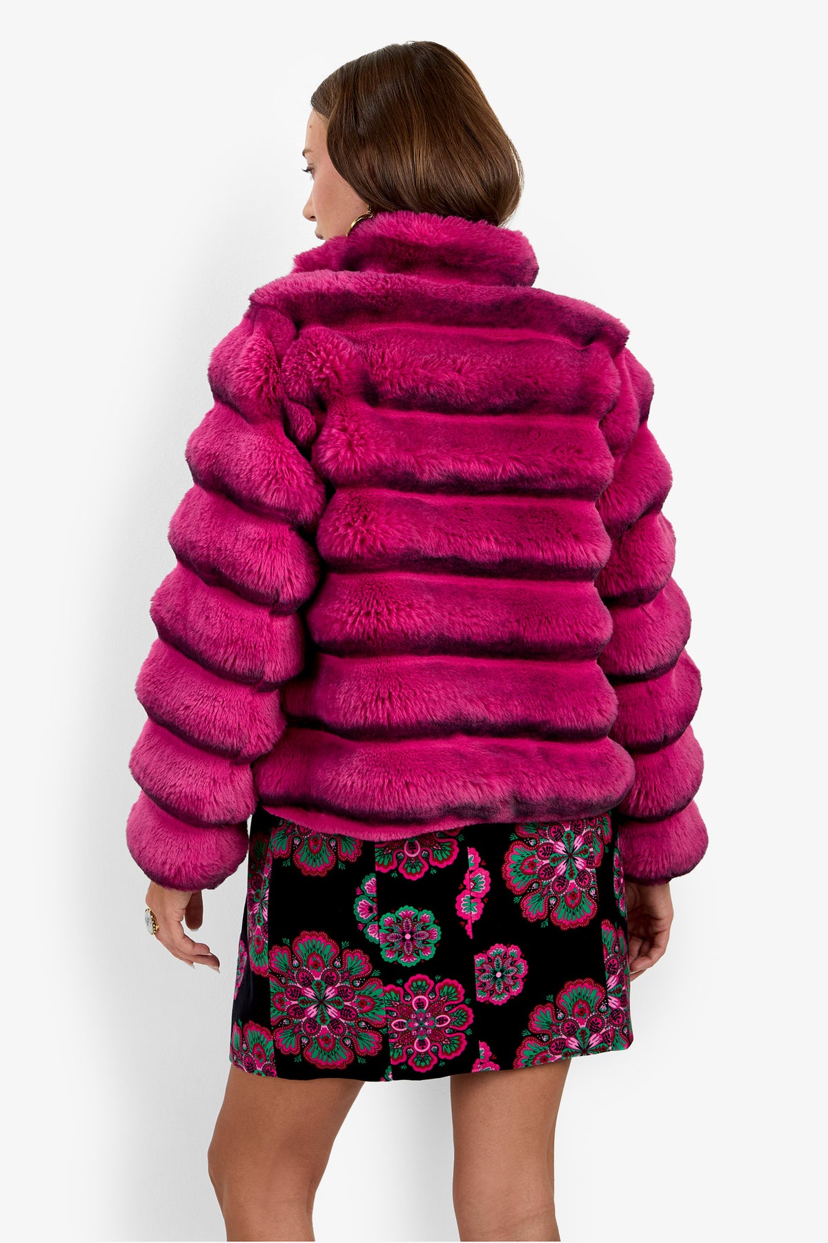 A woman with short brown hair is seen from the back wearing the Mikel Coat, a bright pink faux fur jacket, paired with a black skirt featuring colorful floral patterns.
