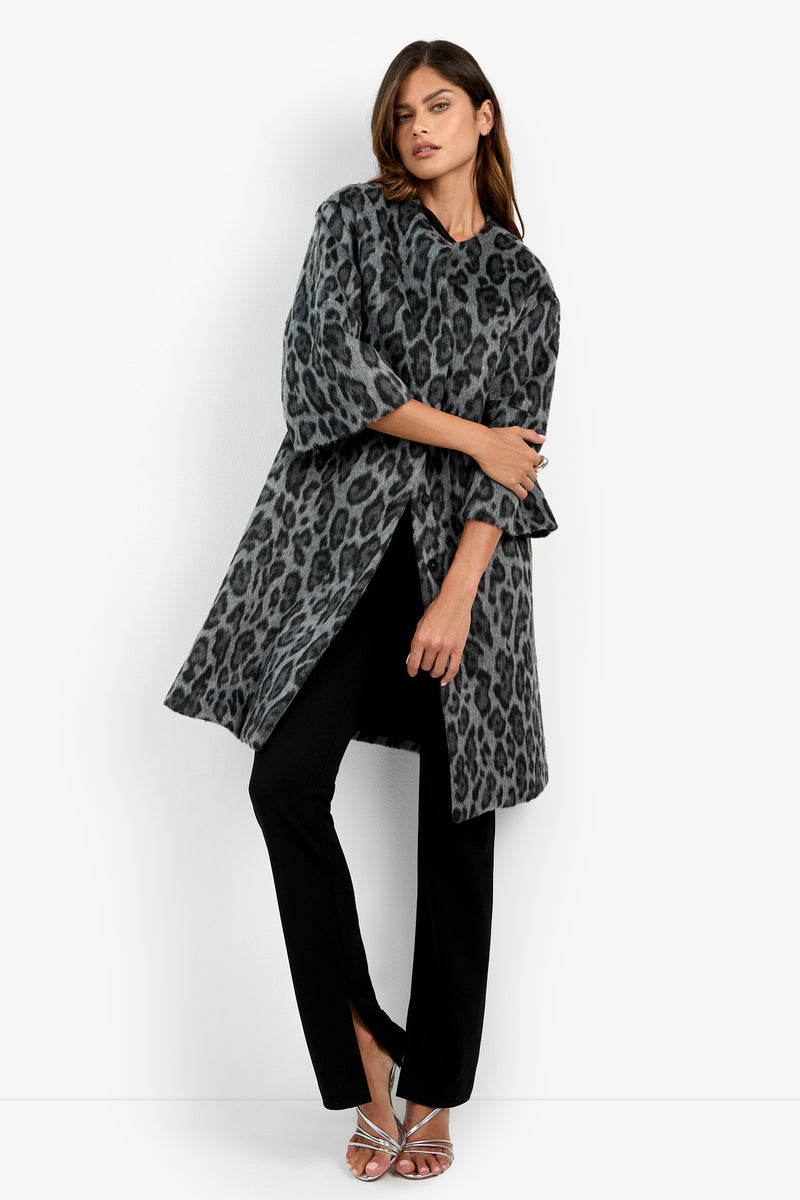 A woman models the Lovely Leopard Coat in grey leopard print, styled with black pants and silver heels. She stands against a plain white background, crossing one leg and resting an arm on her waist.