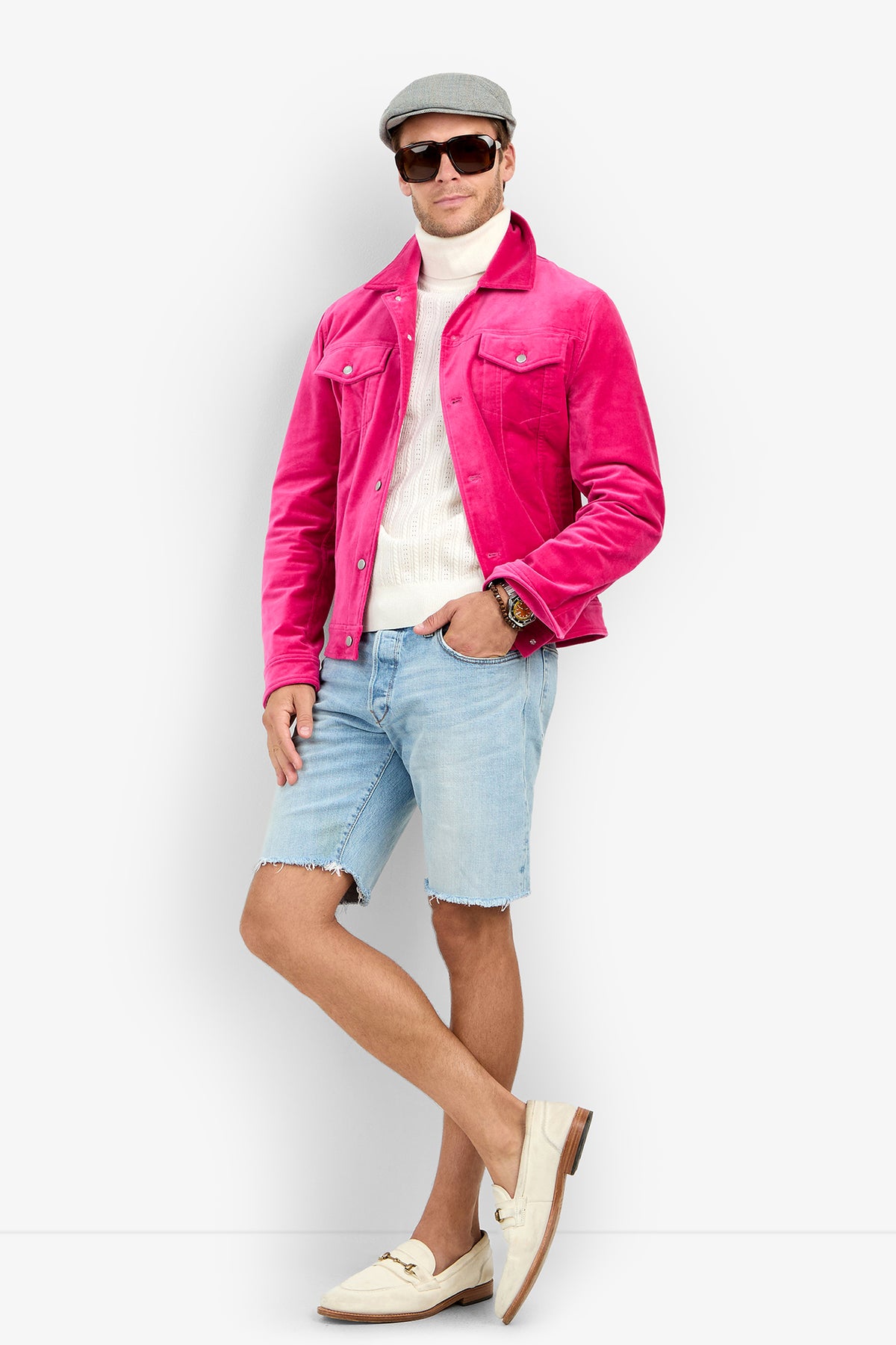 A man wearing a bright pink Angelino Trucker Jacket over a white turtleneck, light-wash denim shorts, a flat cap, and loafers.