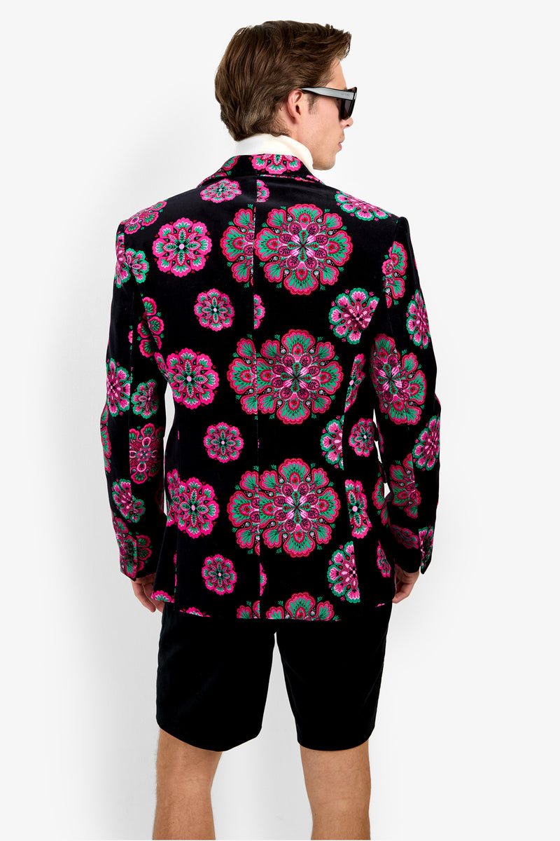 A man wearing sunglasses and black shorts stands facing away, showcasing the back of his Doheny Blazer—a black velvet piece adorned with bright pink and green floral patterns.
