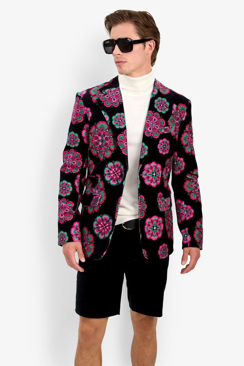 A man stands against a plain white background wearing sunglasses, a white turtleneck, black shorts, and the Doheny Blazer featuring bright pink and turquoise floral patterns.