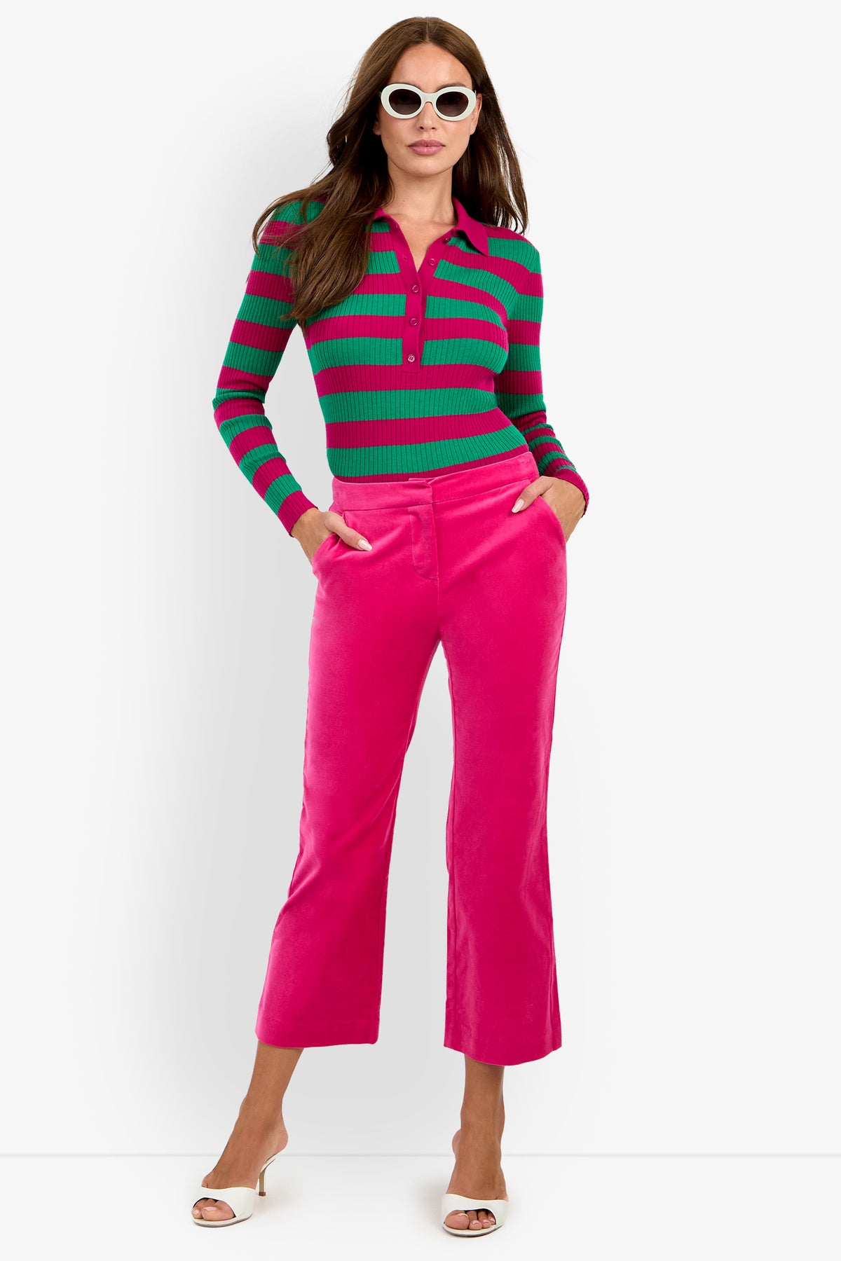 A woman wearing white sunglasses, a green and pink striped long-sleeve sweater, the bright pink Westport Crop pants, and white open-toed heels stands with her hands in her pockets against a white background.