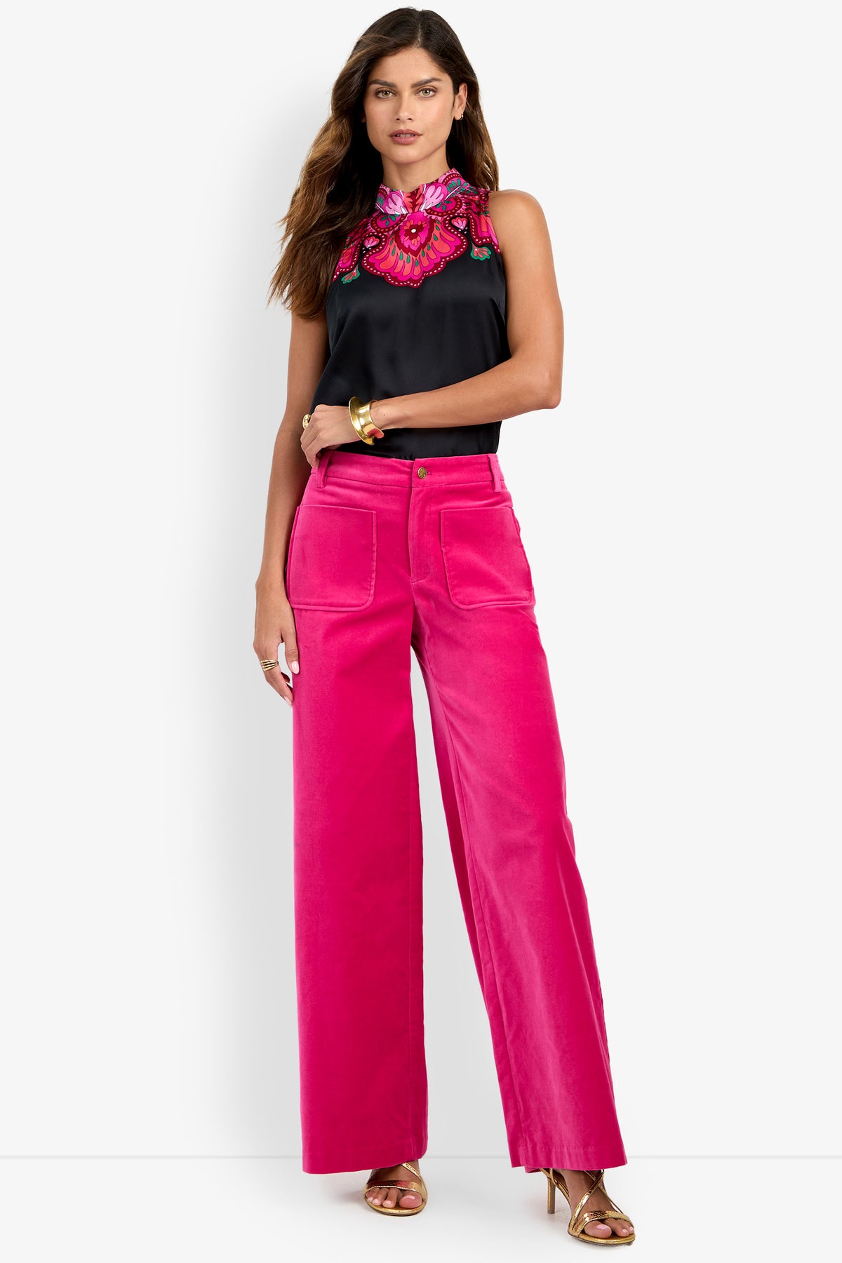 A woman stands against a plain white background wearing bright pink Talamone Pants, a sleeveless black top with pink embroidery, gold bracelets, and gold heels.
