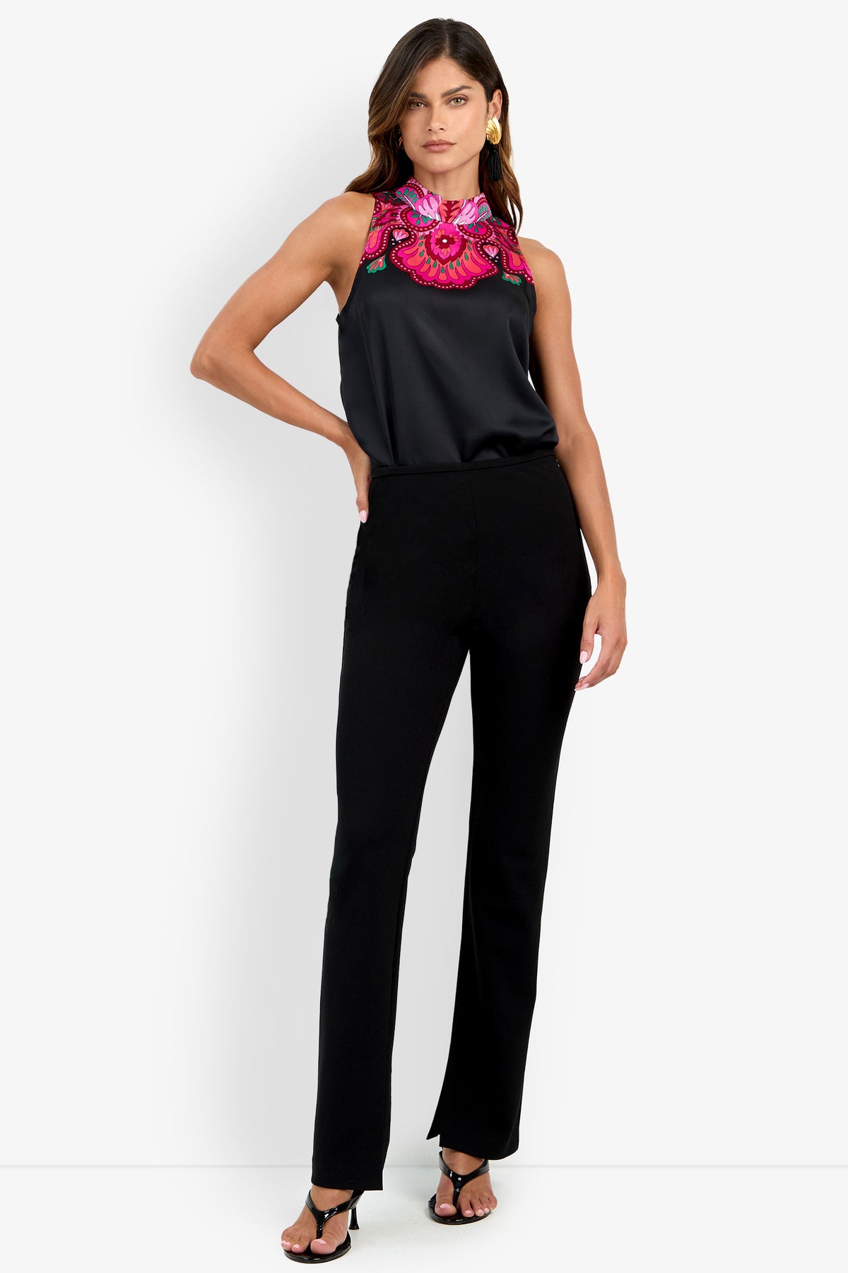 A woman stands against a plain white background with one hand on her hip, wearing the Milena Pant in black, paired with black heels and a sleeveless black top featuring bright pink floral embroidery at the neckline.