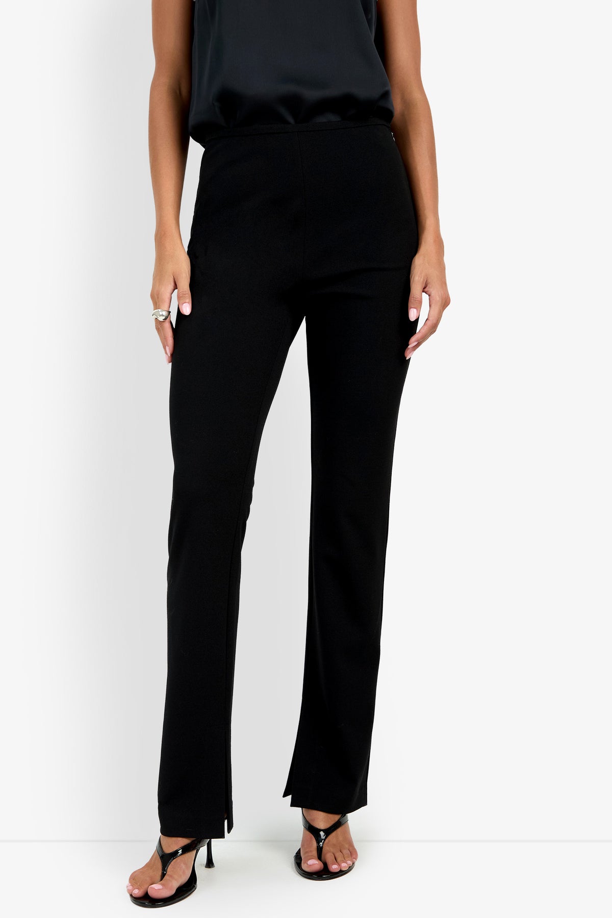A person wearing the Milena Pant in black, paired with a black sleeveless top and black open-toed heels, stands against a plain white background. Only the lower half of the body is visible.