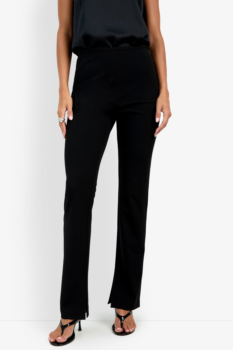 A person wearing the Milena Pant in black, paired with a black sleeveless top and black open-toed heels, stands against a plain white background. Only the lower half of the body is visible.