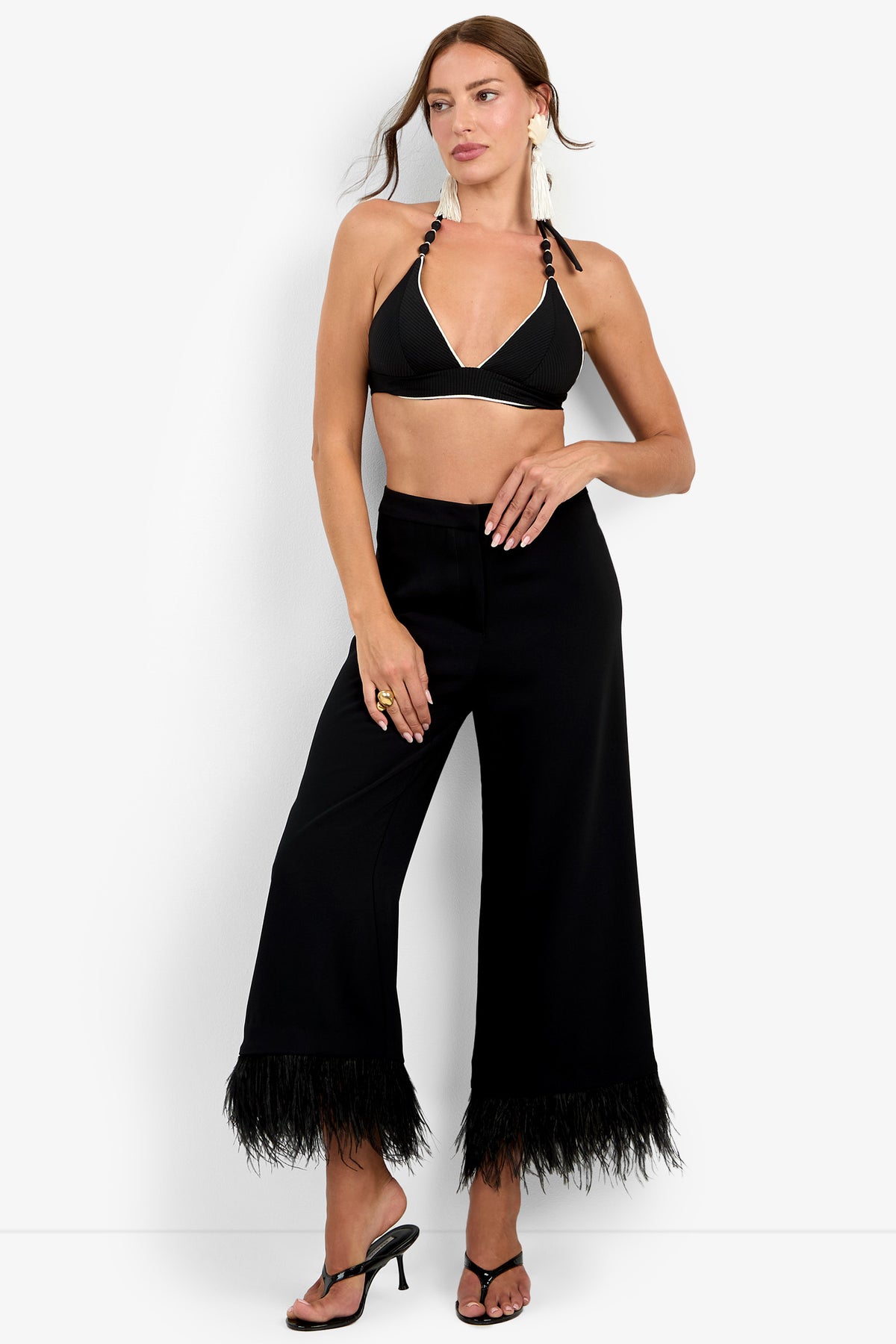 A woman wearing the Amarie Pant—high-waisted black wide-leg pants with feather trim—styled with a black halter bralette, black heeled sandals, large earrings, and a ring, stands against a plain white background.
