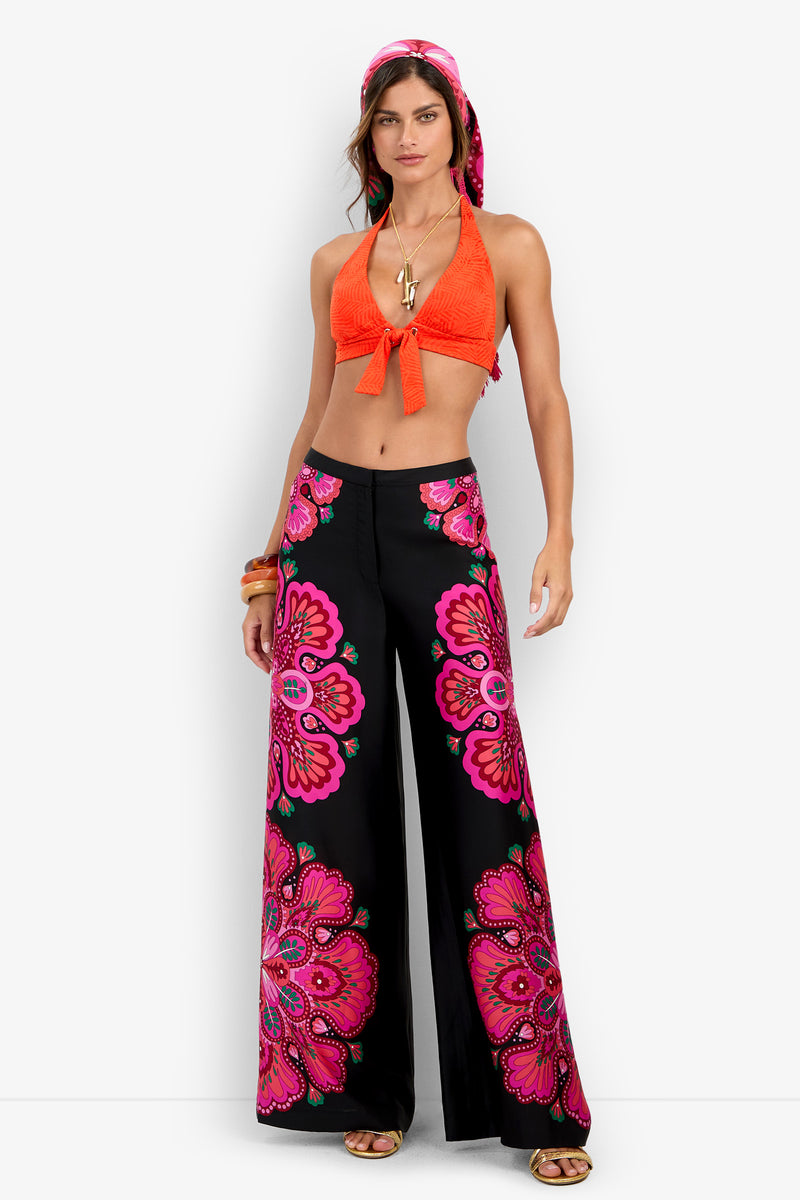 Woman in an orange halter top, the Lanay Pant—black wide-leg pants with large pink floral prints—gold sandals, layered necklaces, bangles, and a pink patterned headscarf standing against a white background.