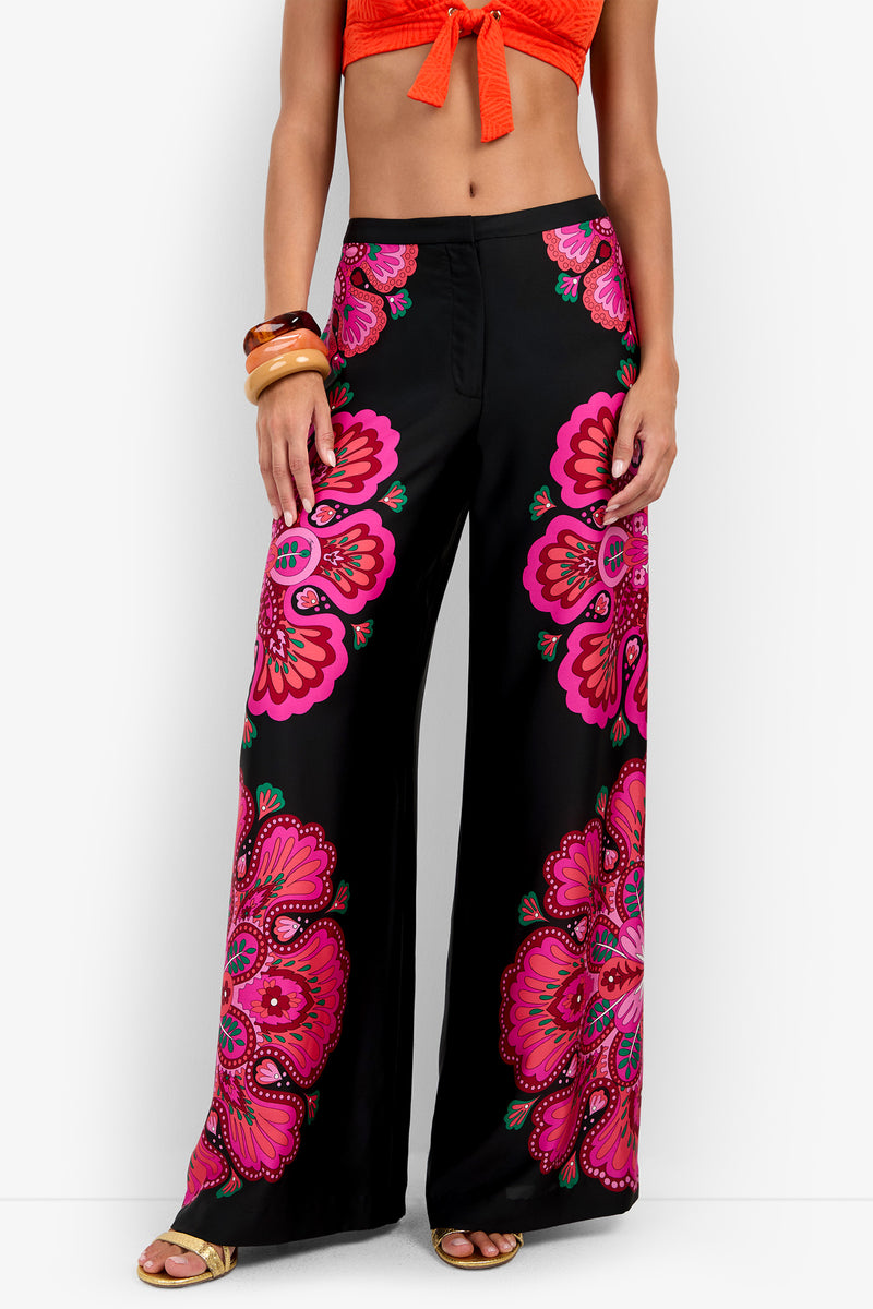A woman models the Lanay Pant—black wide-leg pants adorned with bold pink and red floral patterns—paired with an orange crop top, wooden bangles, and sandals. The photo is cropped from the shoulders down.