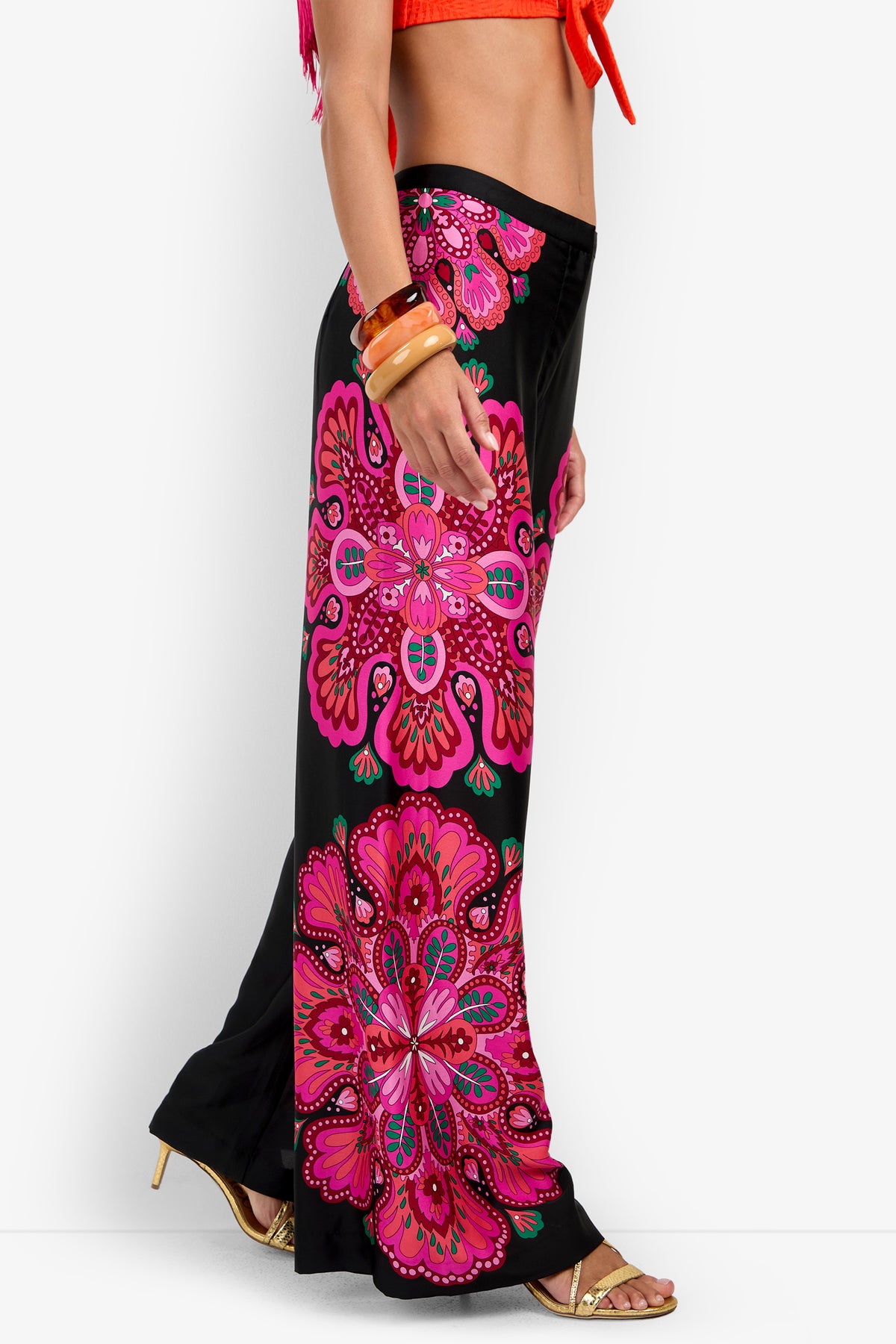 Wearing the Lanay Pant, which features bold pink, red, and green mandala-style patterns on a black background, this individual stands sideways in a bright orange top, gold heels, and chunky orange bracelets.