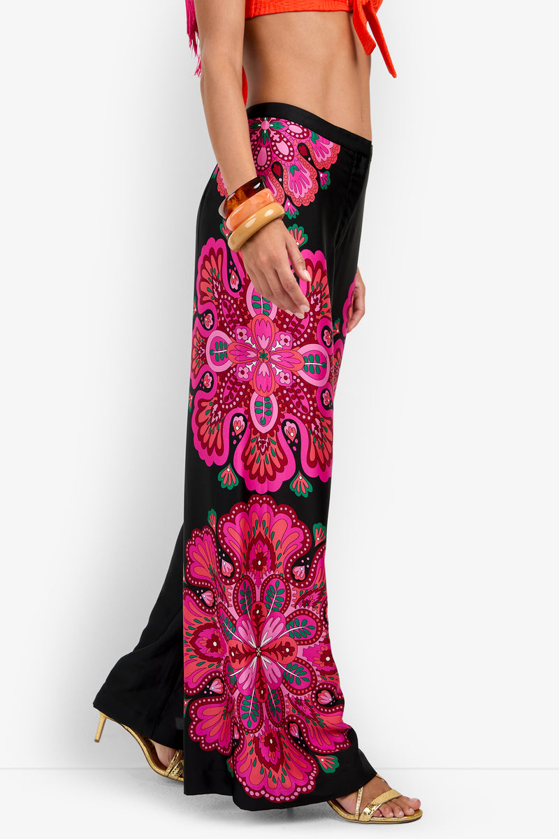 Wearing the Lanay Pant, which features bold pink, red, and green mandala-style patterns on a black background, this individual stands sideways in a bright orange top, gold heels, and chunky orange bracelets.