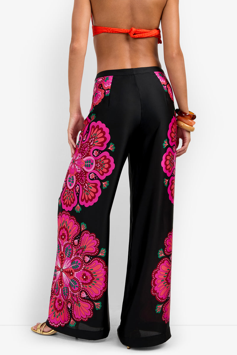 A woman, barefoot and posing with one hand on her hip while facing away from the camera, wears a red bikini top paired with the Lanay Pant—black wide-leg pants featuring bright pink floral patterns.