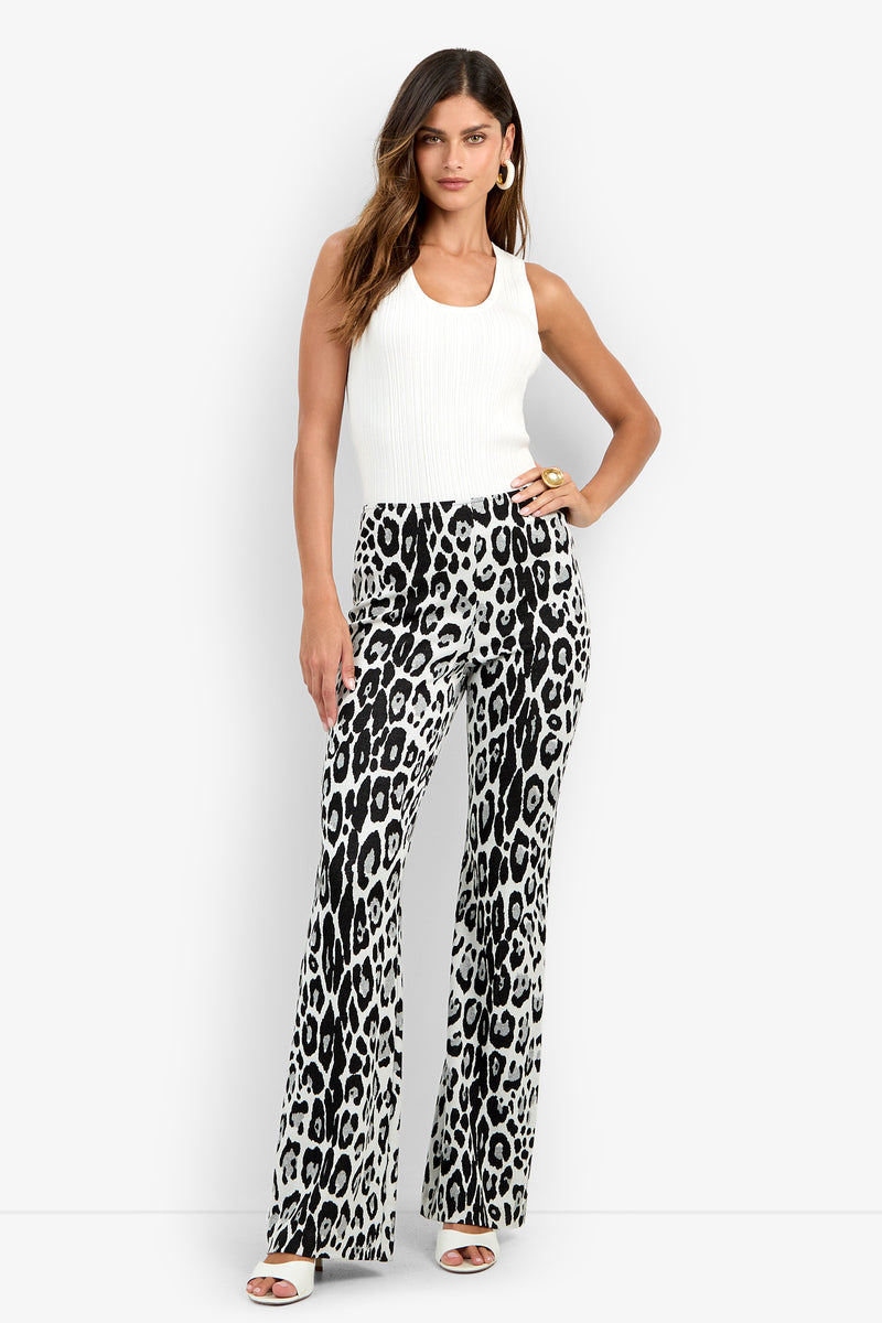 A woman wears the Jacoba Pant in MULTI, featuring a high-waisted, black-and-white leopard print flared silhouette. She pairs it with a sleeveless white top, white sandals, and gold hoop earrings while standing with one hand on her hip.