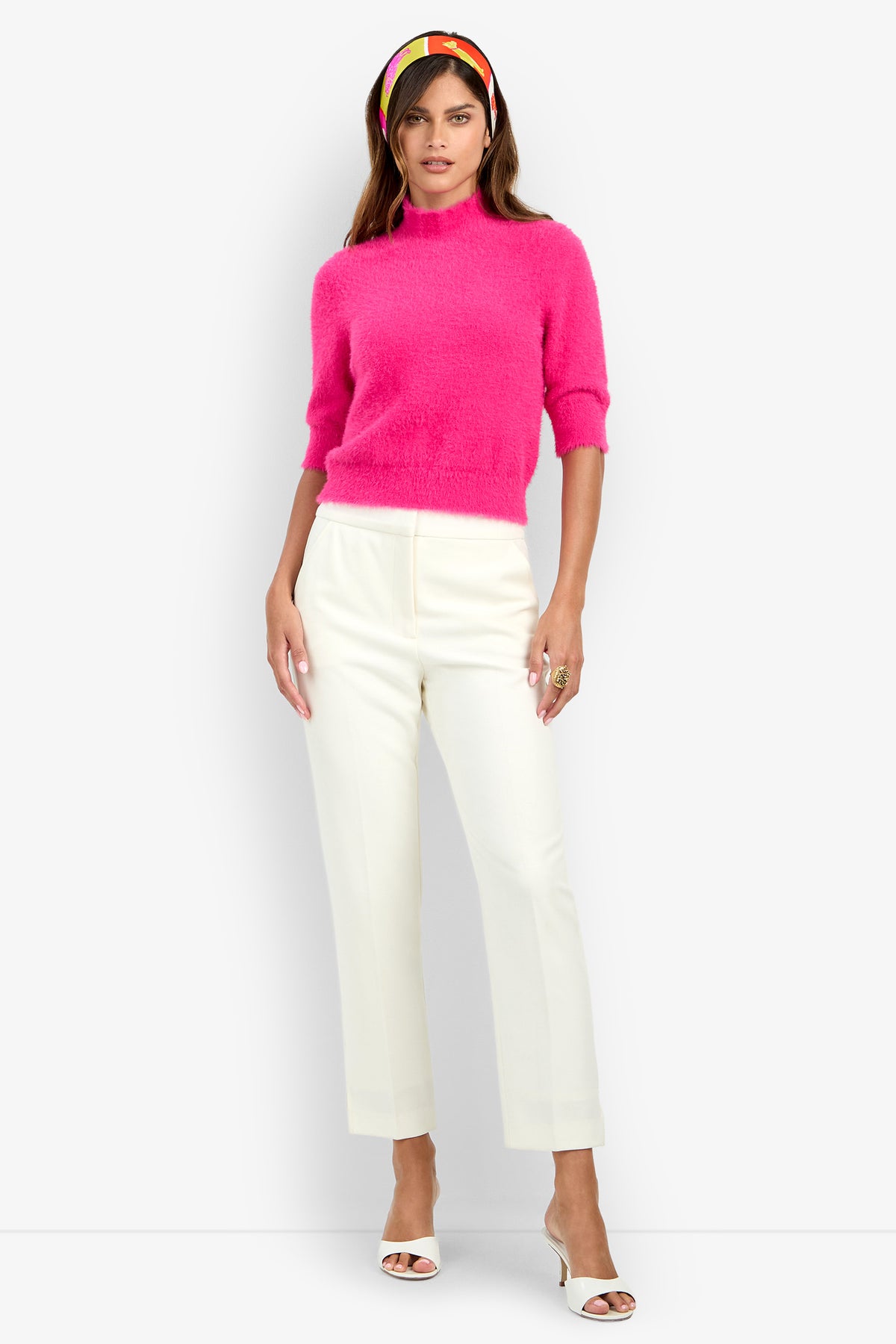 A woman wears the Arno Pant, styled with a bright pink fuzzy sweater, white heeled sandals, and a colorful headband as she stands against a plain light gray background.