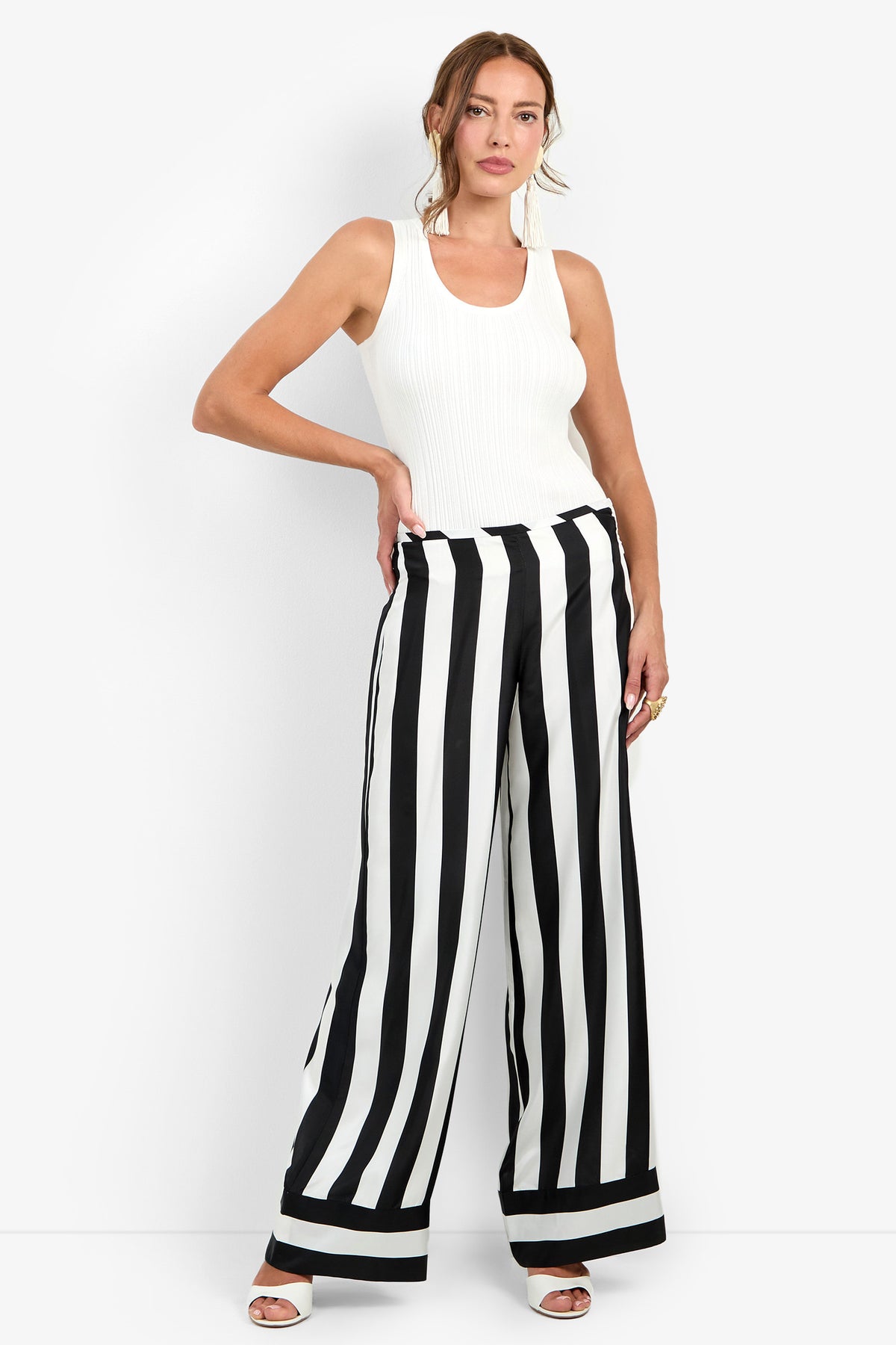 A woman stands against a white background, wearing a sleeveless white top, the Khiara Pant in BLACK/WINTER WHITE—a high-waisted, wide-leg pant with black-and-white vertical stripes—and white heels.