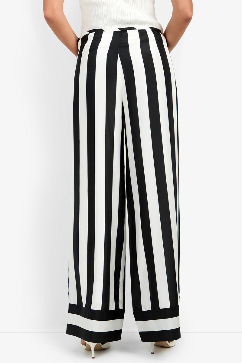 A person seen from the back wearing the Khiara Pant—high-waisted, wide-leg trousers featuring bold vertical black and white stripes—paired with light heels and a tucked-in white top.