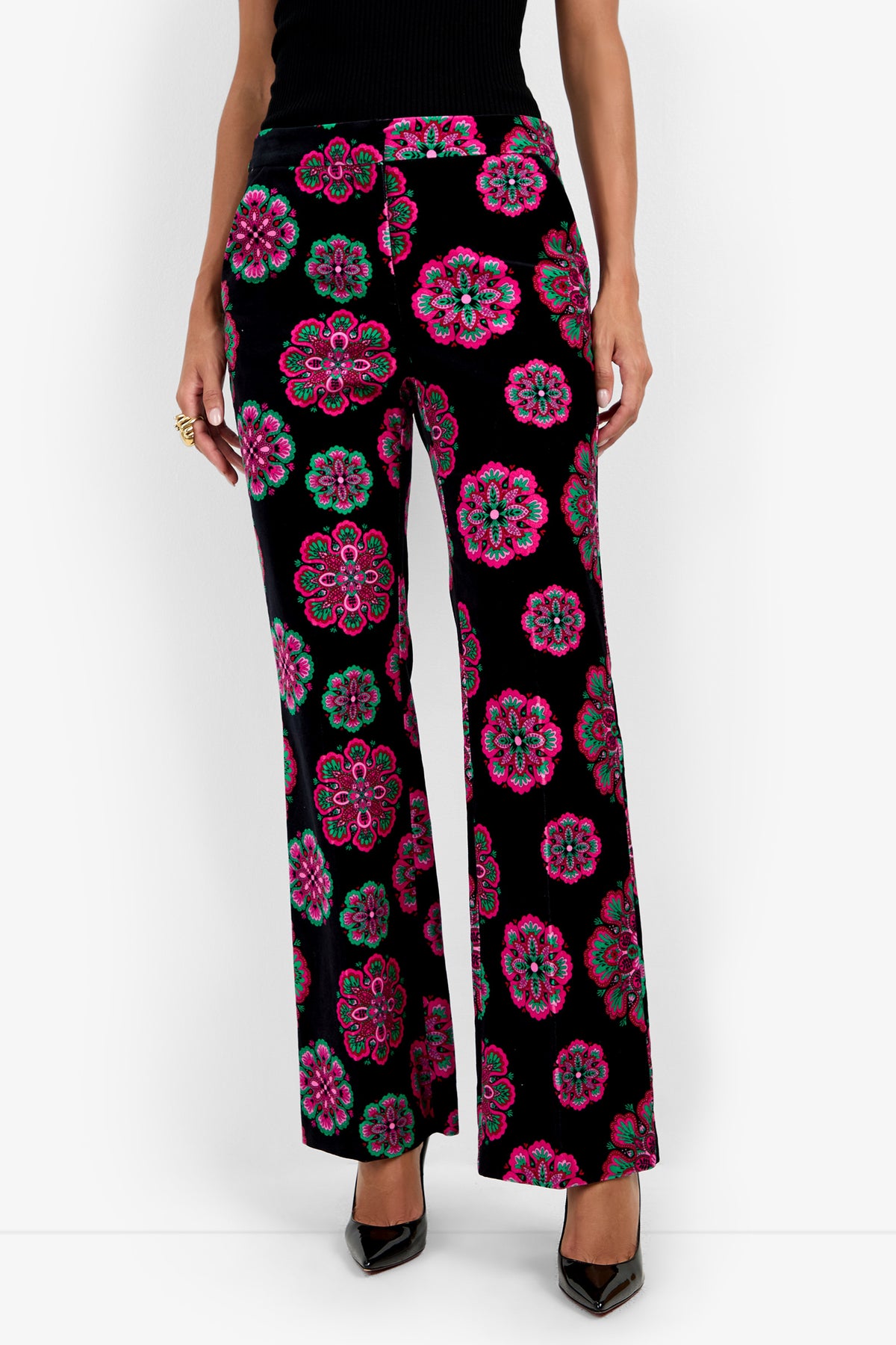 A person wearing the Westport Pant, featuring bright pink, green, and blue floral patterns on a black background, paired with a sleeveless black top and black pointed-toe heels. Only the lower body is visible.