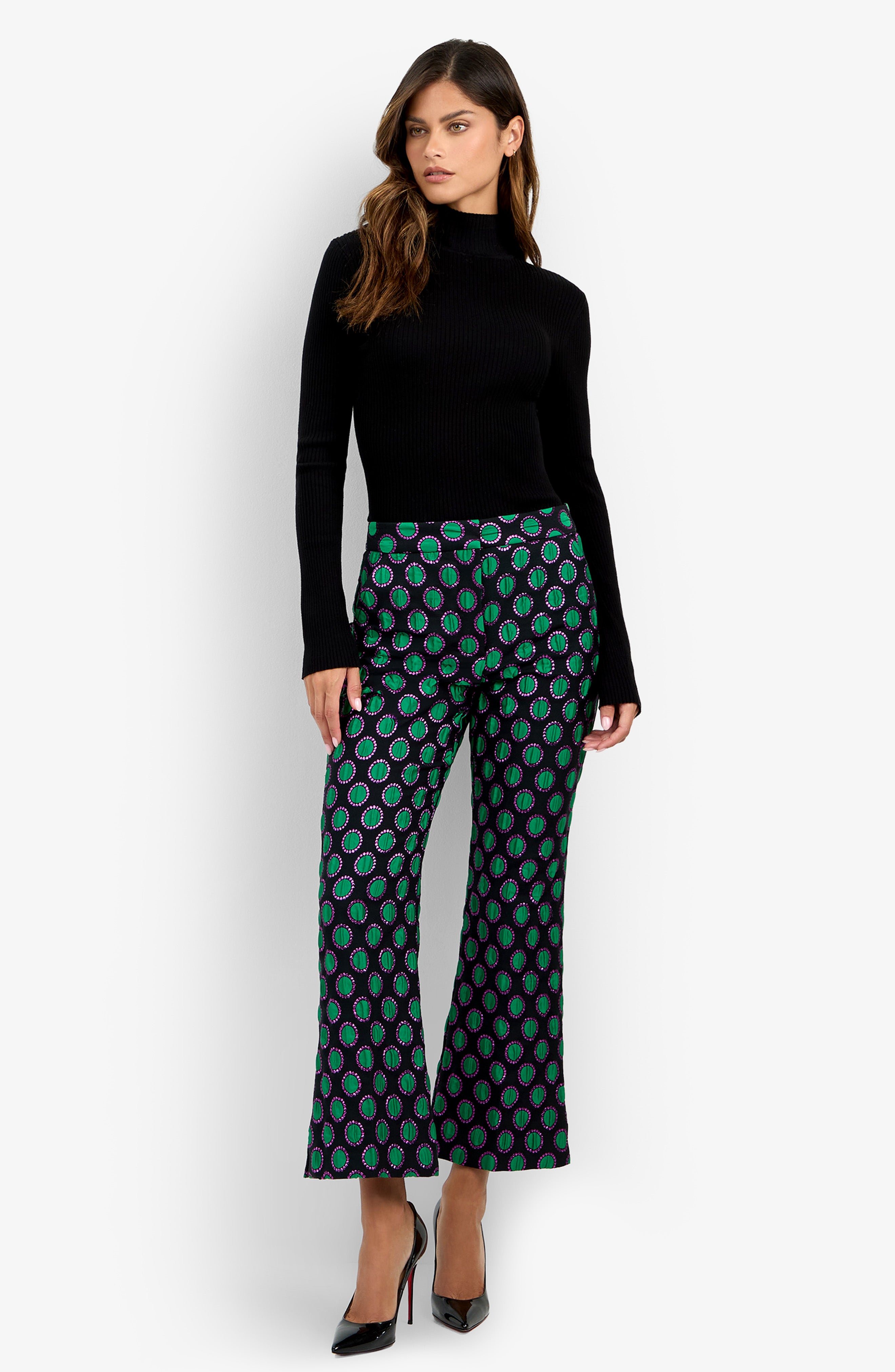 A woman stands against a plain white background wearing a black turtleneck sweater, black high heels, and the Allisan Pant—wide-leg pants featuring a green and purple pattern.