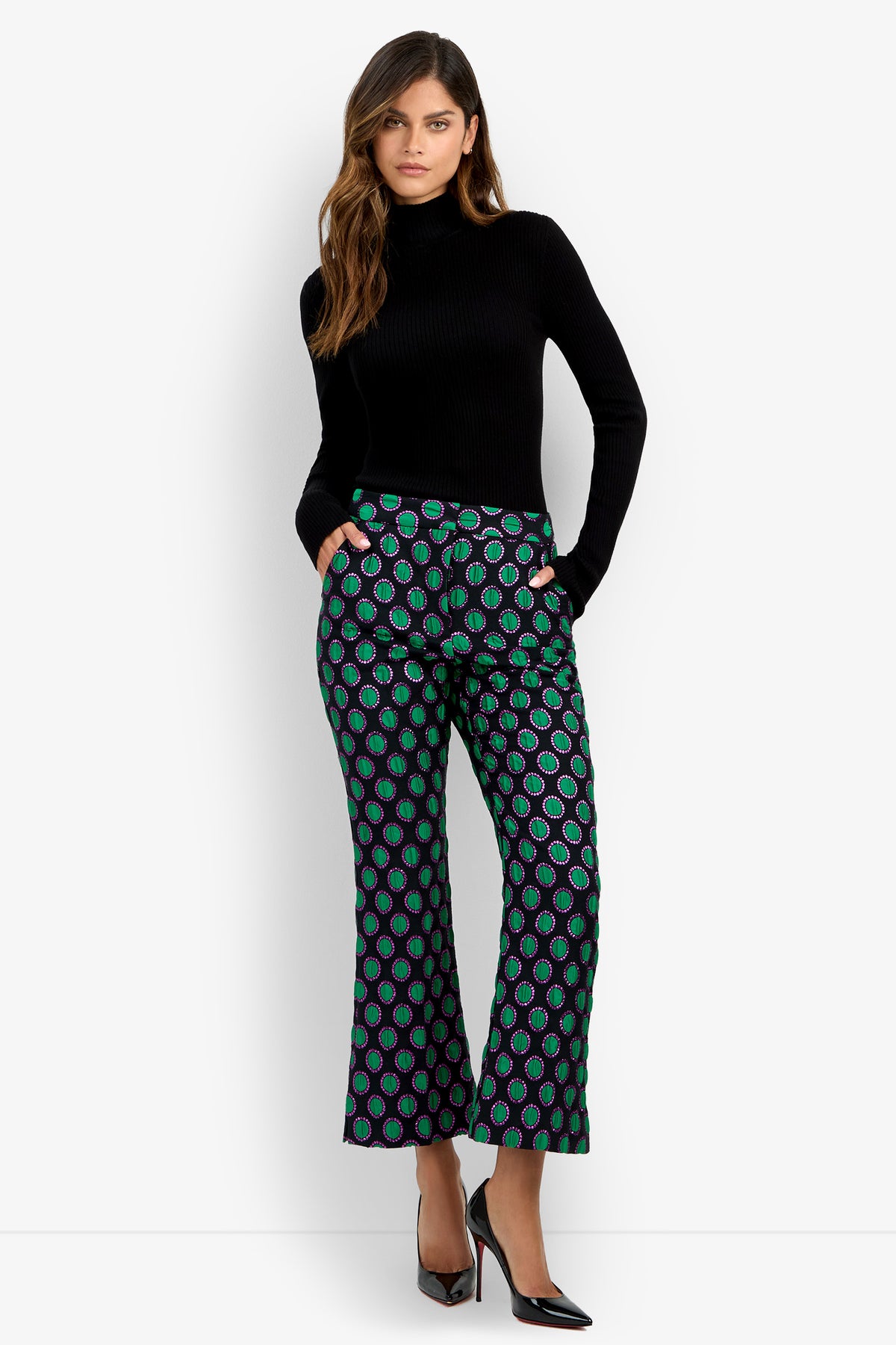 A woman stands with one hand in her pocket against a plain white background, wearing the Allisan Pant—a pair of green patterned wide-leg pants—styled with a black turtleneck sweater and black heels.