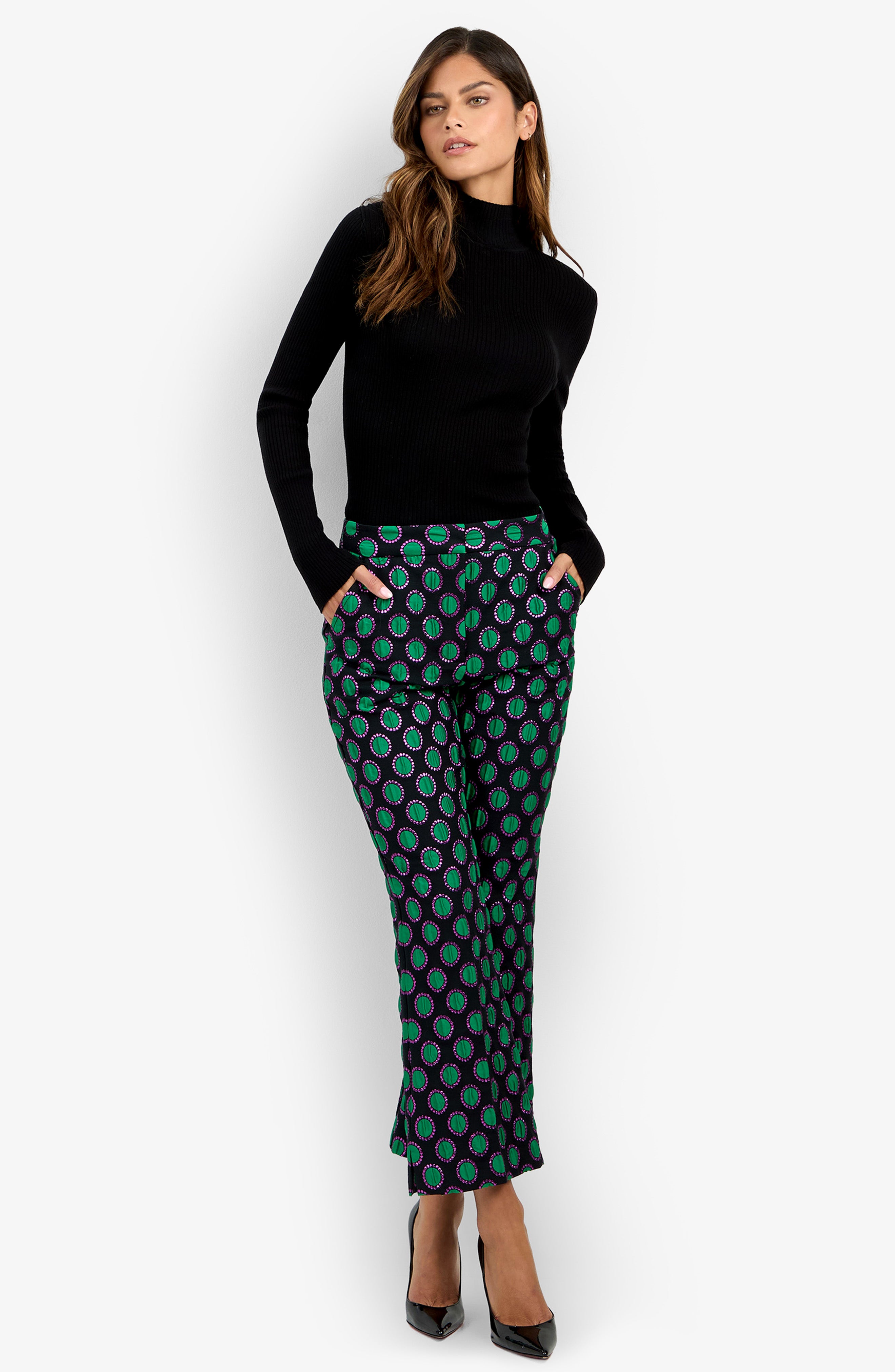 A woman stands with her hands in her pockets, wearing a black long-sleeve top, the patterned green and purple Allisan Pant, and black high heels against a plain white background.