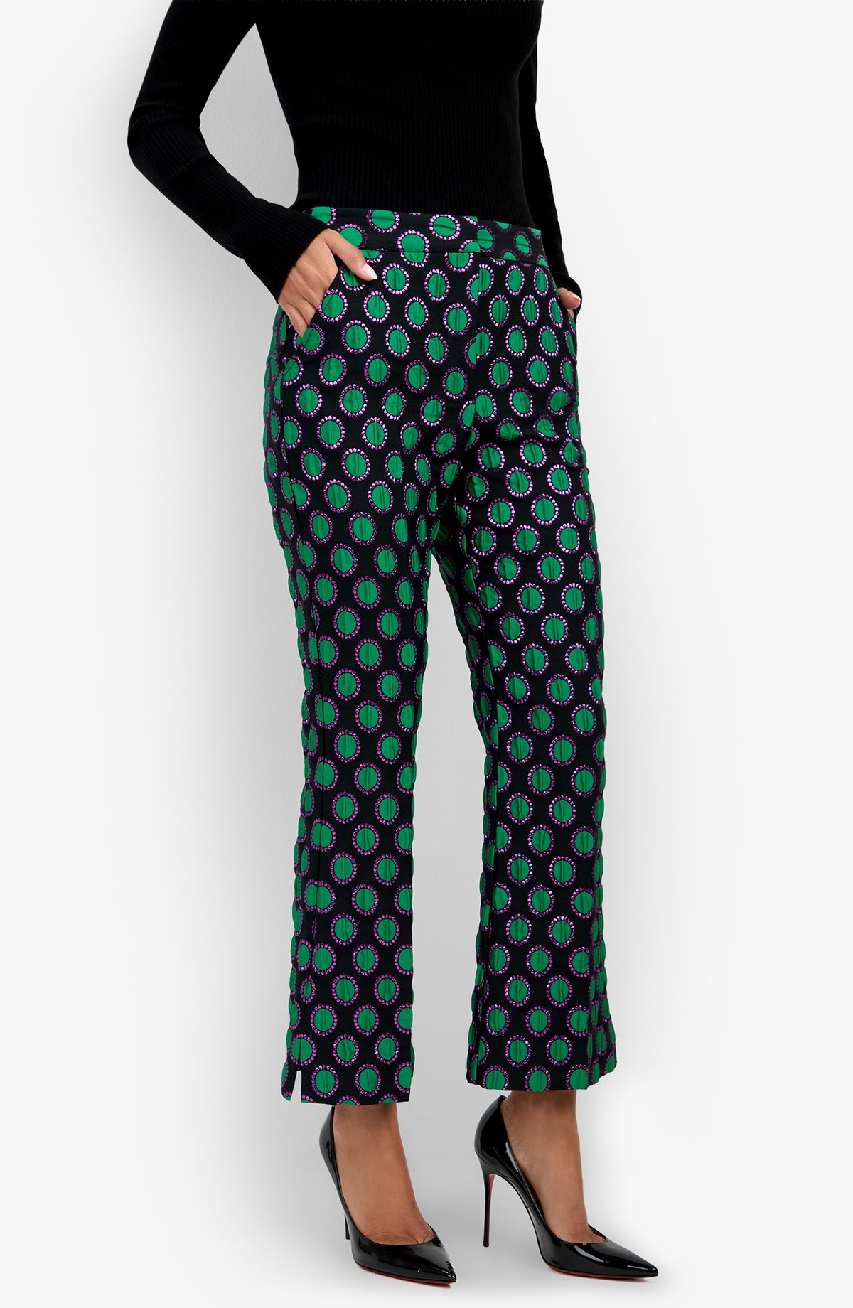 A woman in a black turtleneck wears the Allisan Pant—patterned in green, purple, and blue—with her hands in her pockets and paired with black high-heeled shoes, standing against a white background.