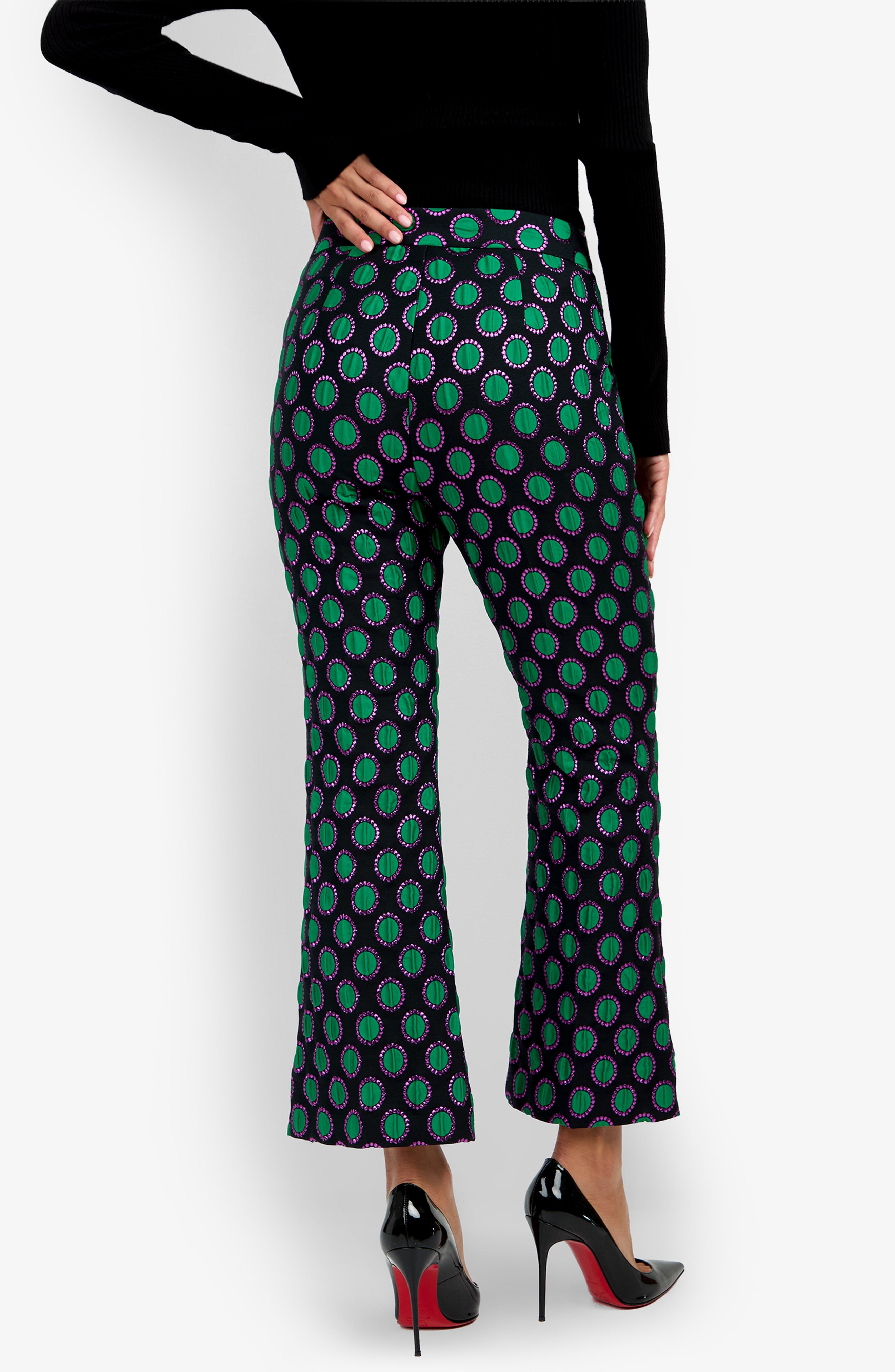 Person wearing the Allisan Pant, a pair of high-waisted, flared pants with a green and purple circular pattern, styled with black high heels and a black long-sleeve top, seen from the back.