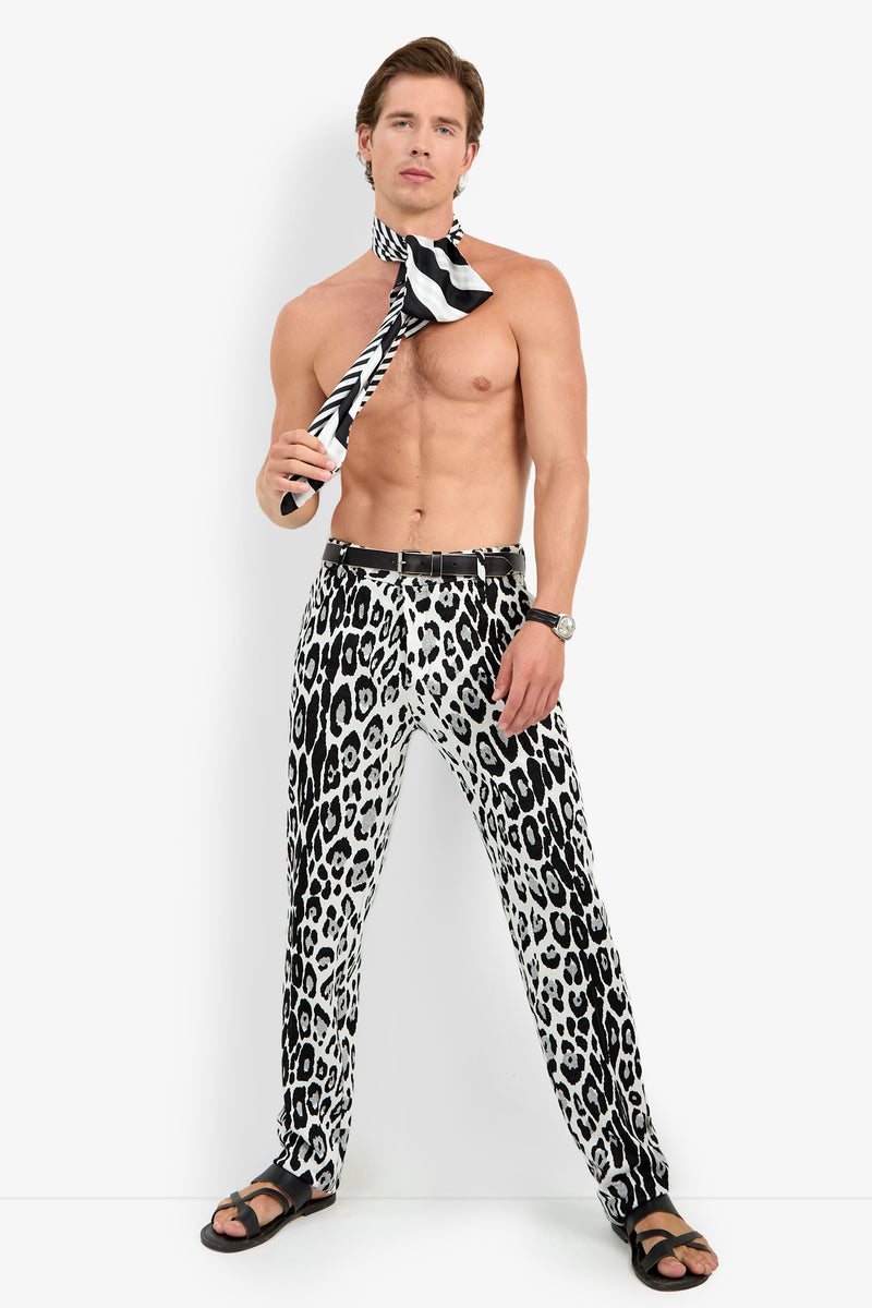 Shirtless man standing, wearing the Clyde Slim Trouser in black-and-white animal print, a matching striped scarf around his neck, sandals, and a watch, posing against a plain white background.