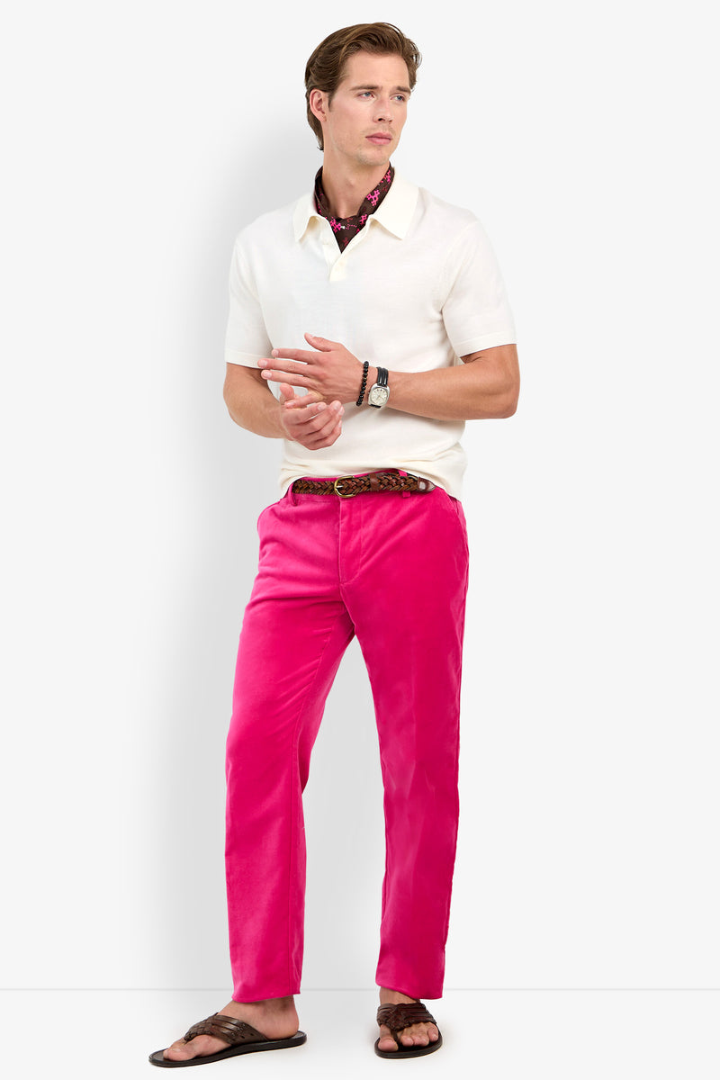 A man stands wearing a white short-sleeve polo, the Clyde Slim Trouser in bright pink, a belt, brown sandals, a patterned scarf, and wrist accessories against a plain white background.