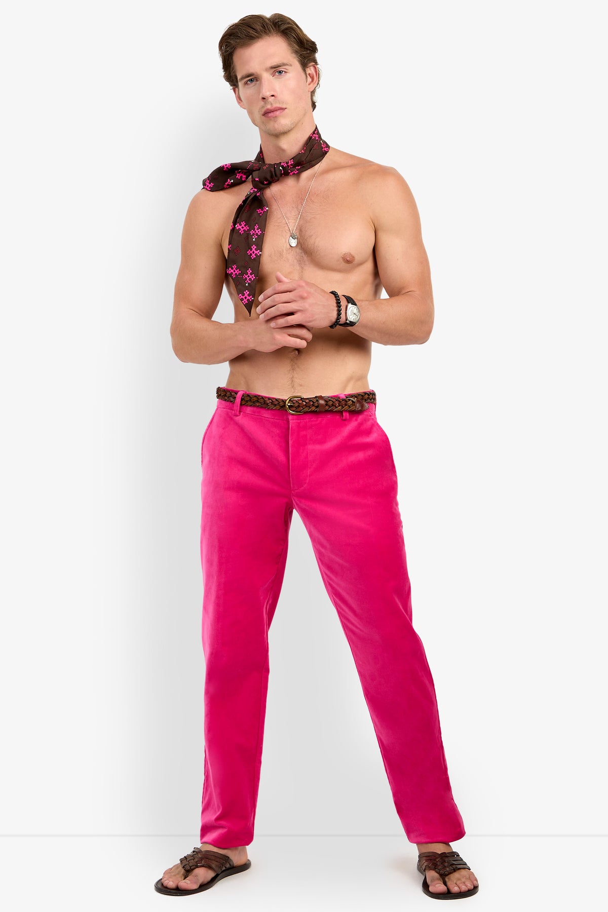 A shirtless man wears the Clyde Slim Trouser in bright pink, paired with a brown patterned scarf tied around his neck, a watch, a necklace, and flip-flops. He stands in a relaxed pose against a plain white background.