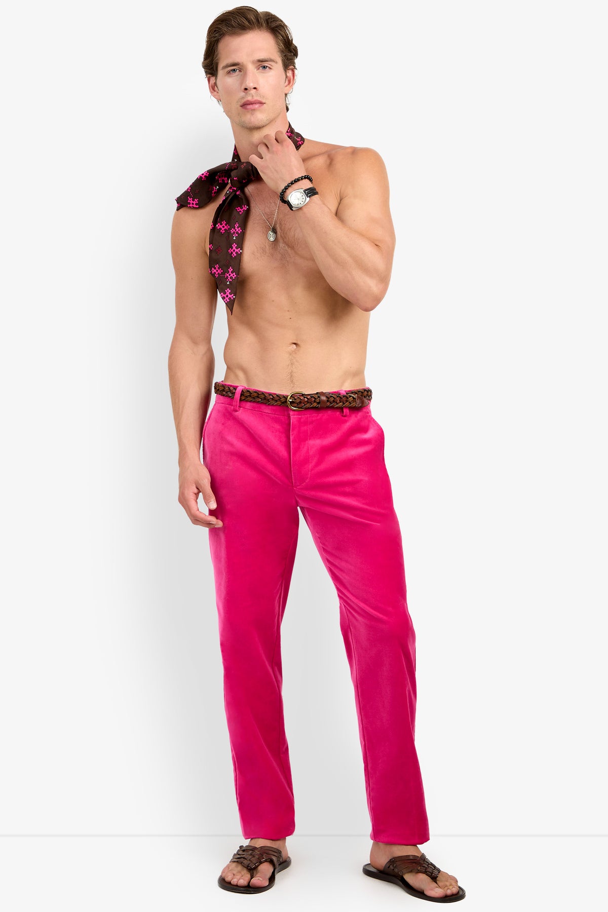 A shirtless man models the Clyde Slim Trouser in bright pink, paired with a patterned belt, sandals, a watch, and a necklace. He holds a black scarf with pink stars over his shoulder while standing against a plain light background.