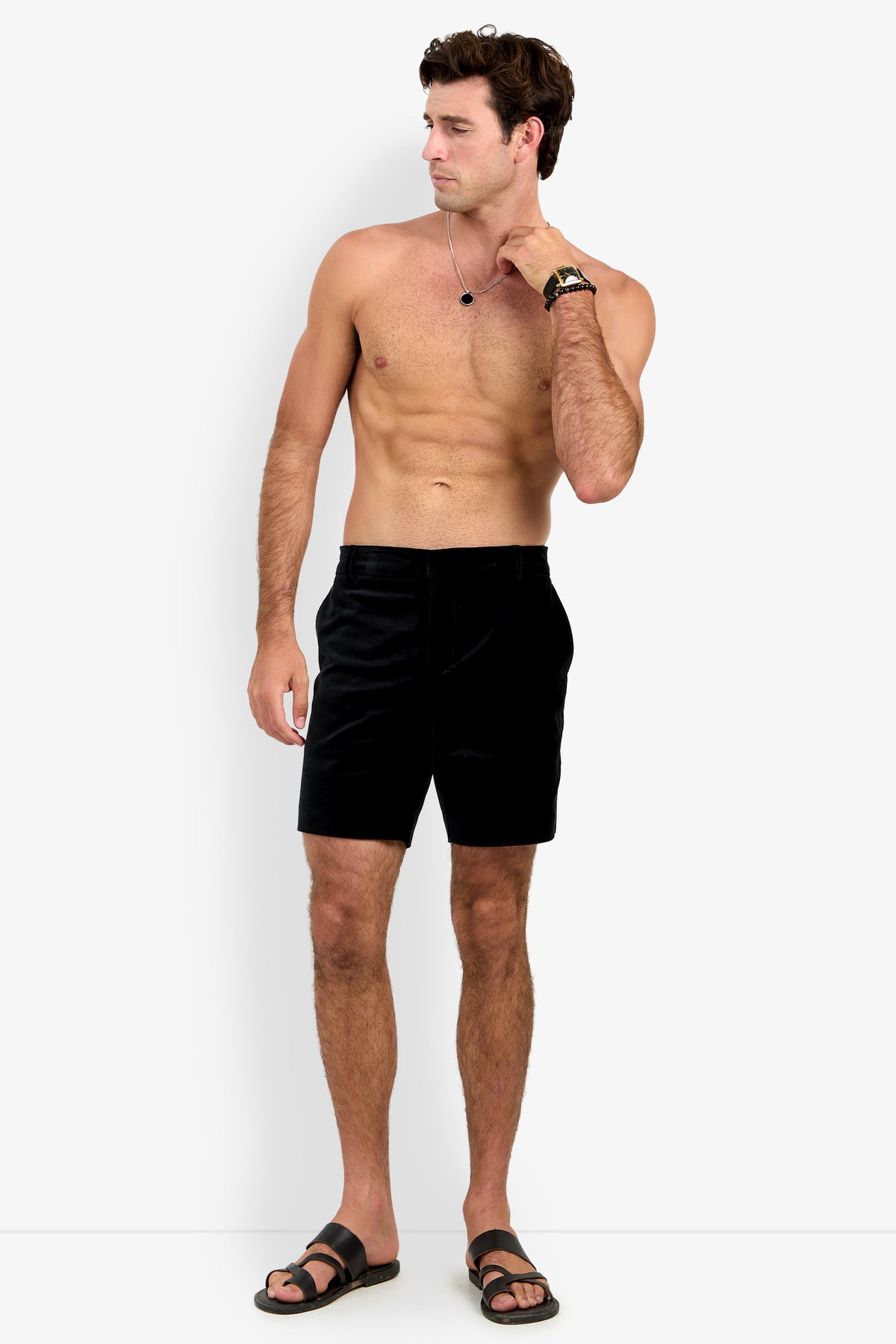 A shirtless man stands against a white background wearing the LAWRENCE SHORT in black, paired with black sandals, a watch, and a necklace. His left hand touches his neck as he looks to his left.