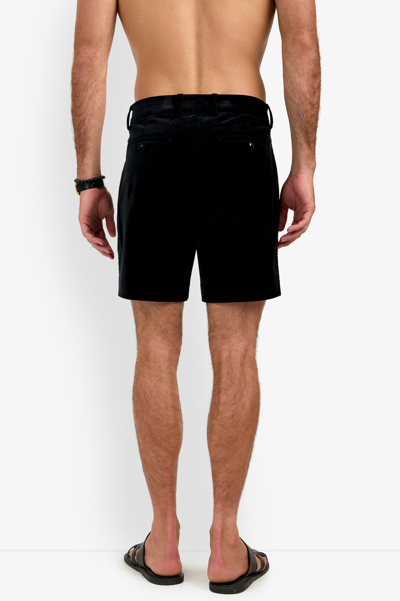 A shirtless man stands with his back to the camera against a plain white background, wearing the LAWRENCE SHORT in black, paired with black flip-flops and a black bracelet.