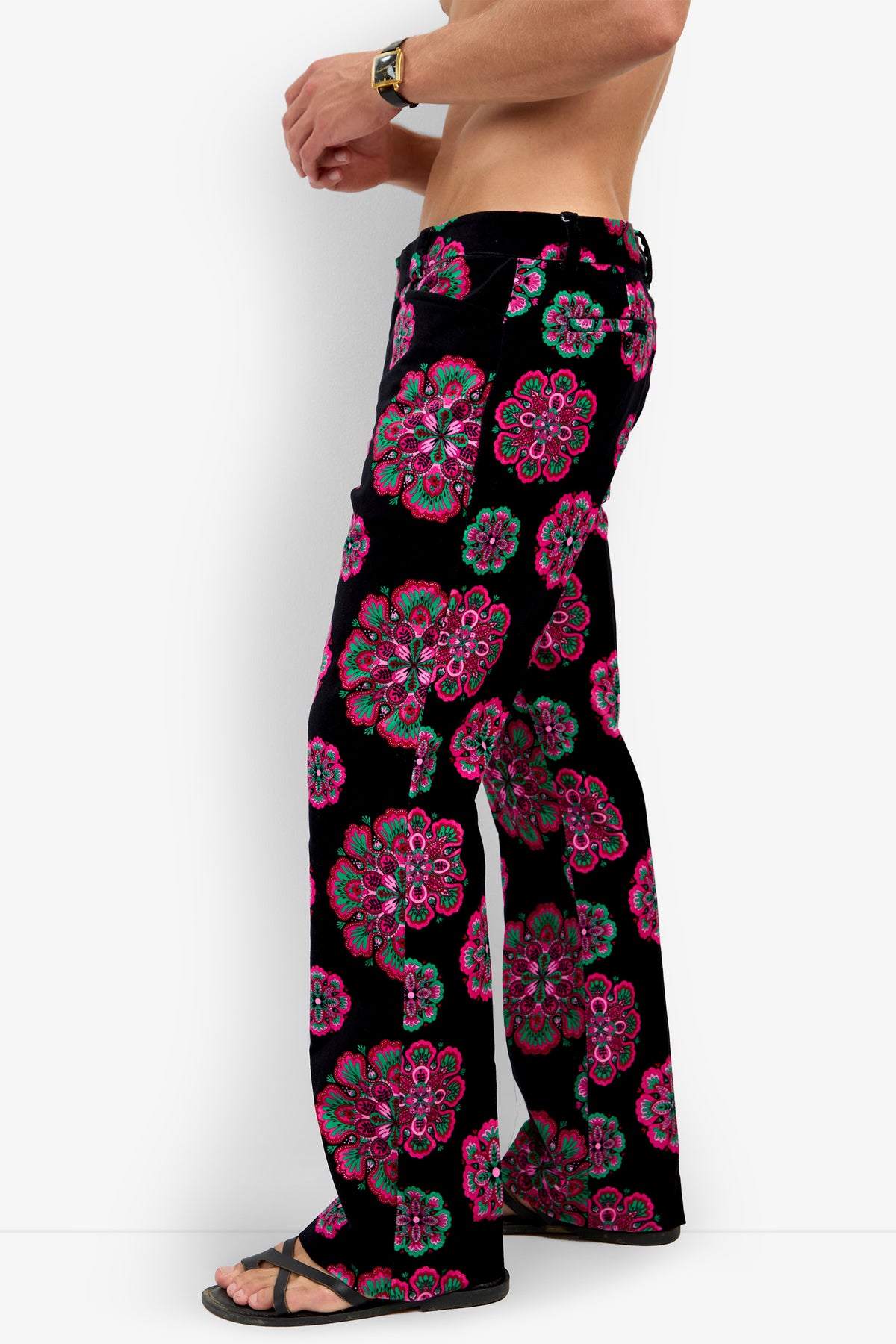 A shirtless person wearing the Alejandro Bootleg Trouser—black flared pants featuring bright pink and green floral patterns—paired with black sandals and a watch, shown from the side against a plain white background.