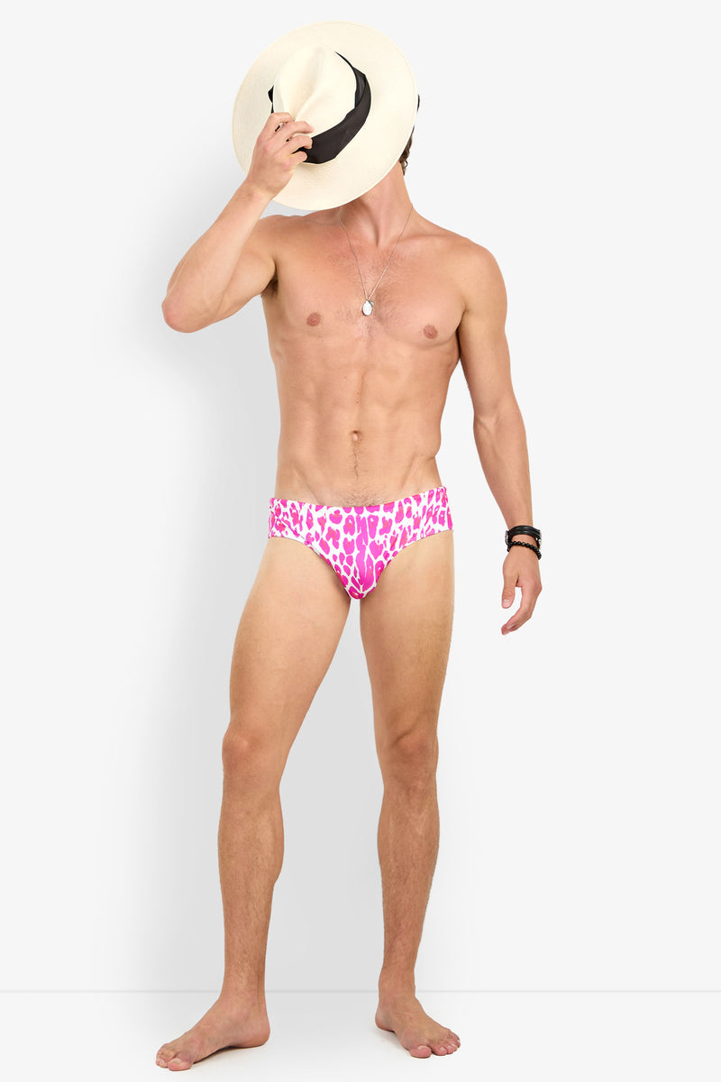 A shirtless person models the BONDI SWIM BRIEF in a pink animal print, covering their face with a wide-brimmed hat. They accessorize with a necklace and bracelets, standing against a plain white background.