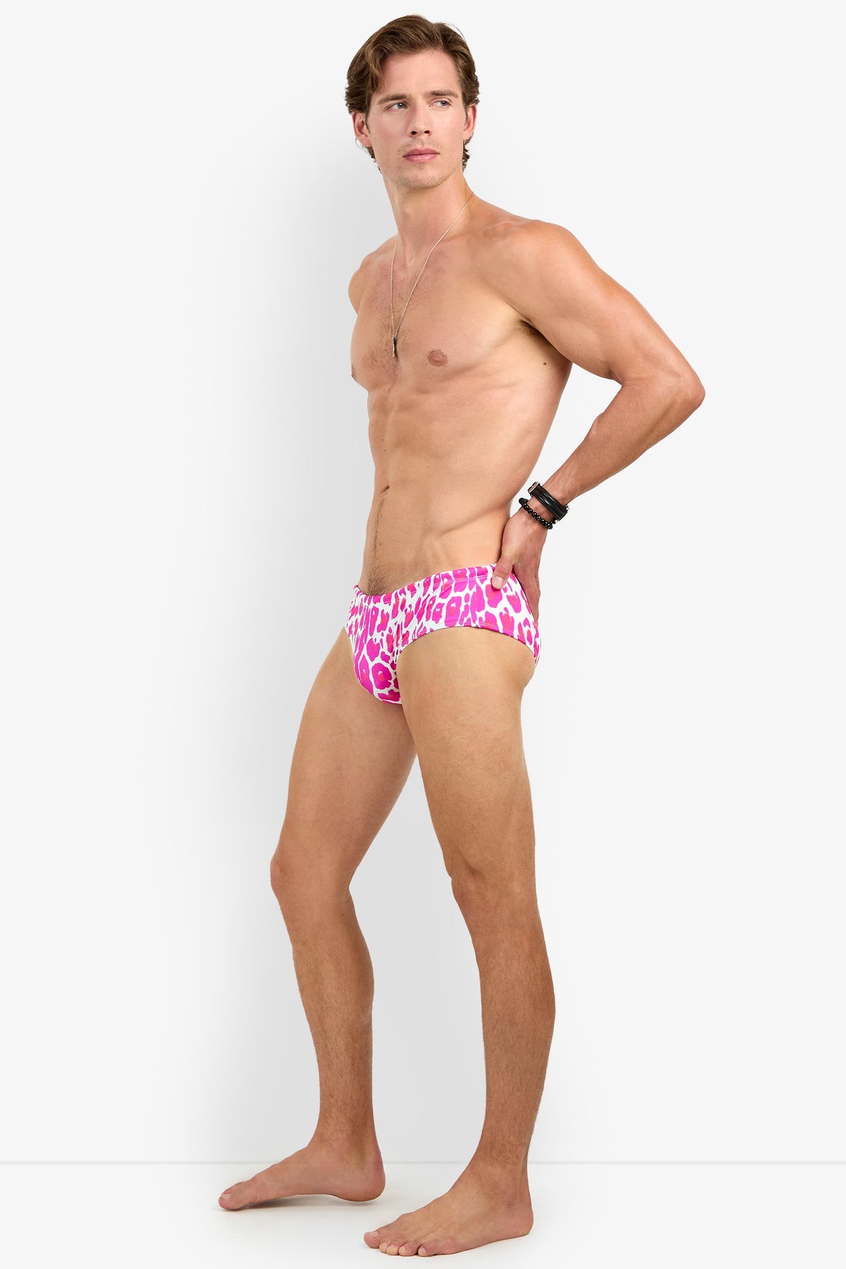 A shirtless man models the BONDI SWIM BRIEF—featuring a pink and white animal print—standing barefoot against a plain white background, gazing to the side with one hand on his hip.