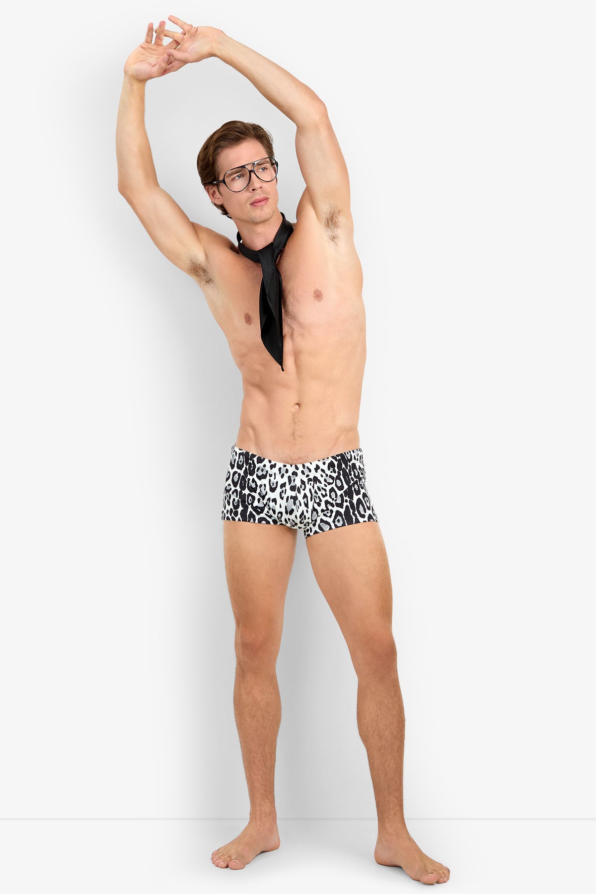 A man wearing glasses, a black scarf, and MONTEGO BAY SWIM TRUNK stands barefoot and stretches with his arms raised to one side.