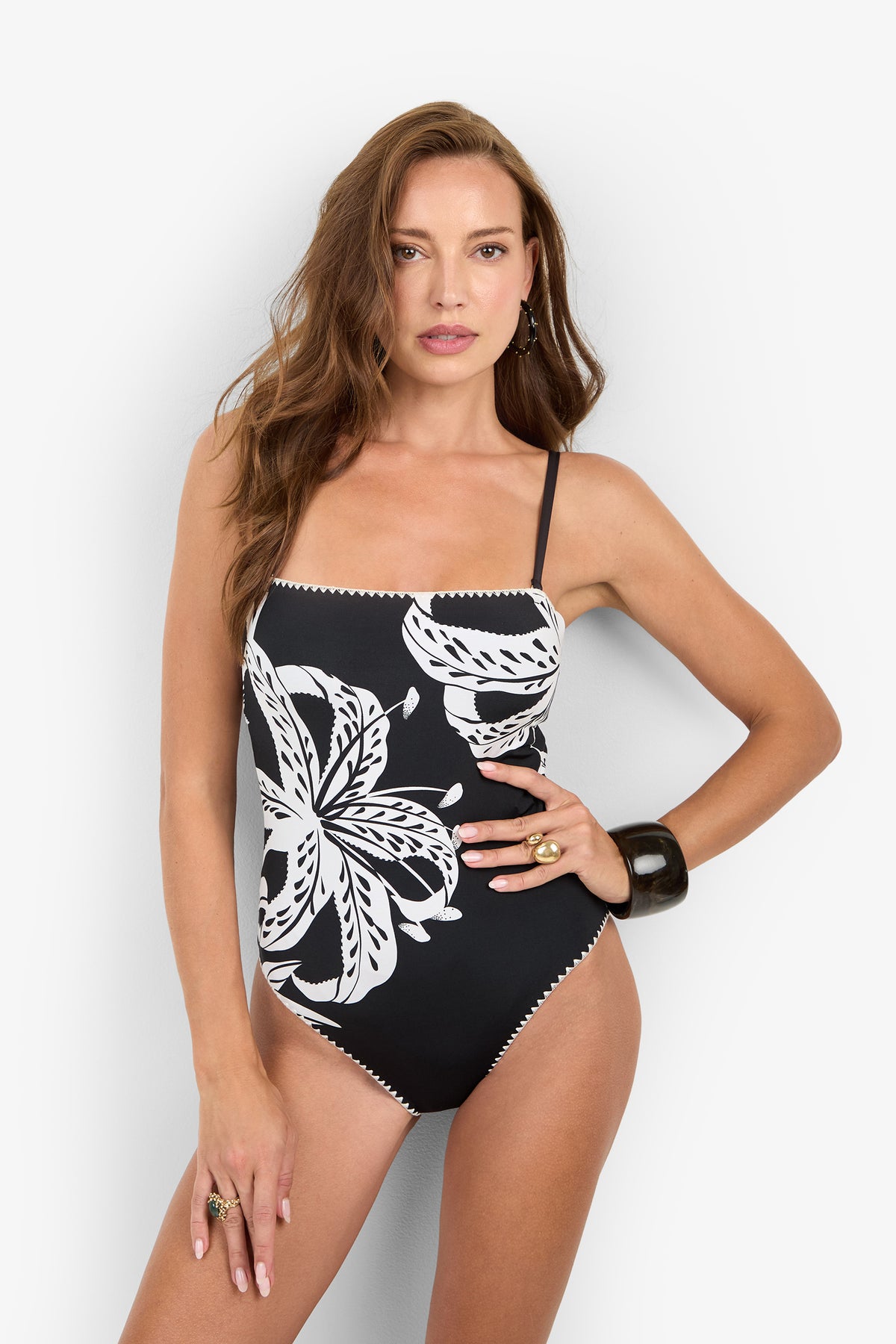 A woman models the GOLDEN HOUR BLOOM BANDEAU ONE PIECE, a black bandeau swimsuit featuring a large white floral print, spaghetti straps, and a chic floral design.